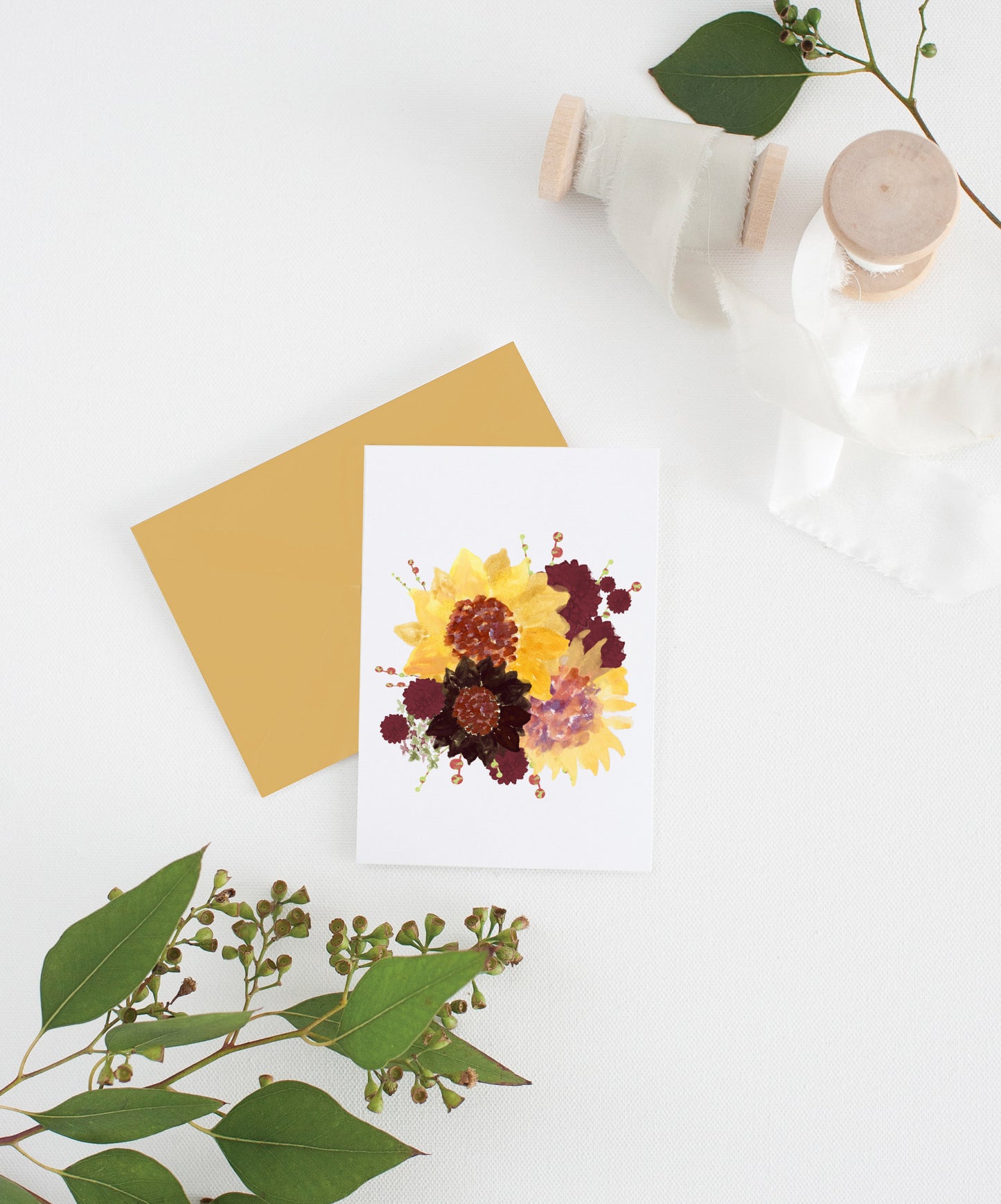 Greeting card with colorful envelope laying on a white surface with green leaves and cotton ribbon wound on wooden spools.