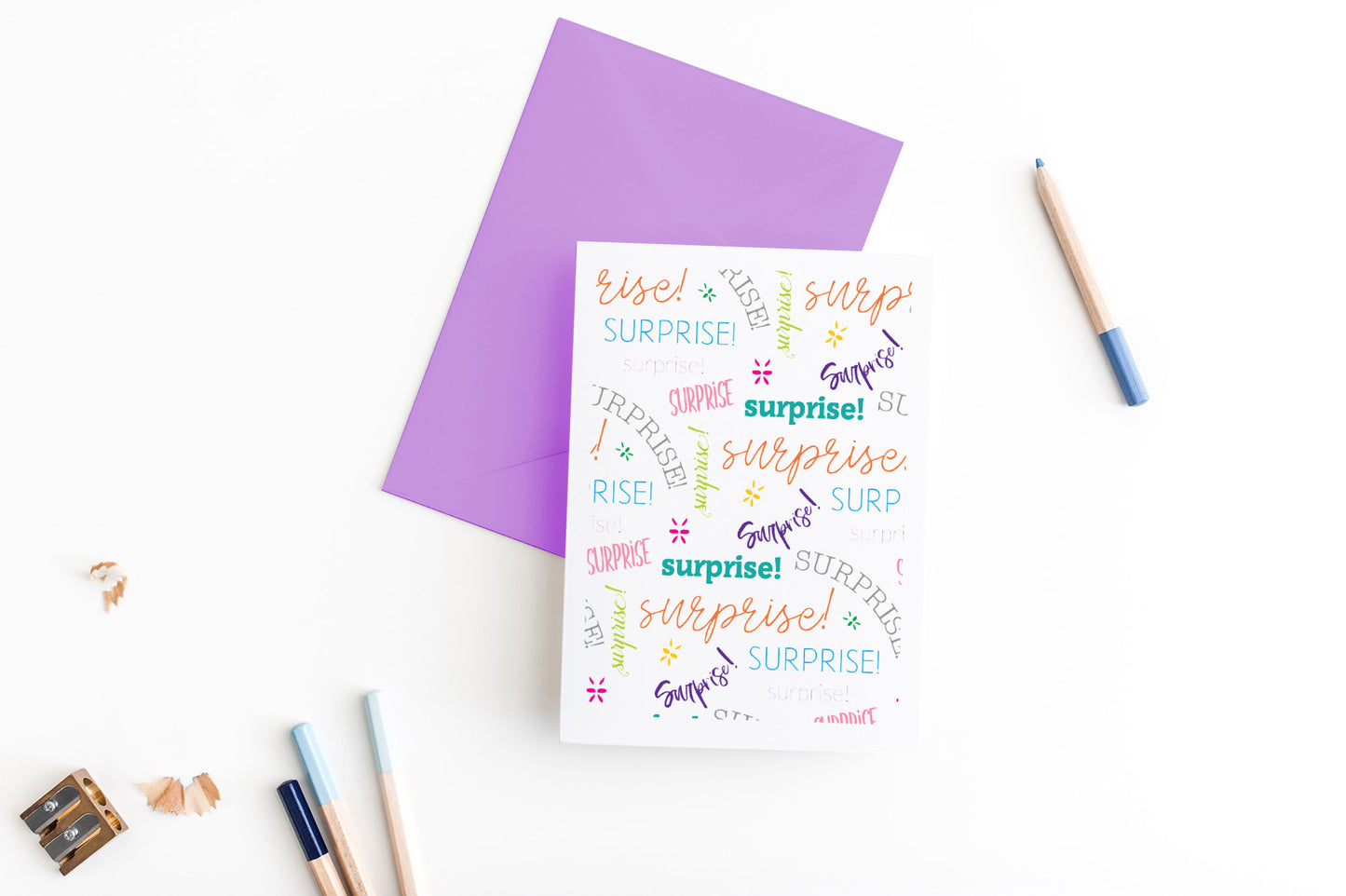 Greeting card, seen from above, with a colored, square flap envelope, pencils, pencil sharpener and shavings on a white background.