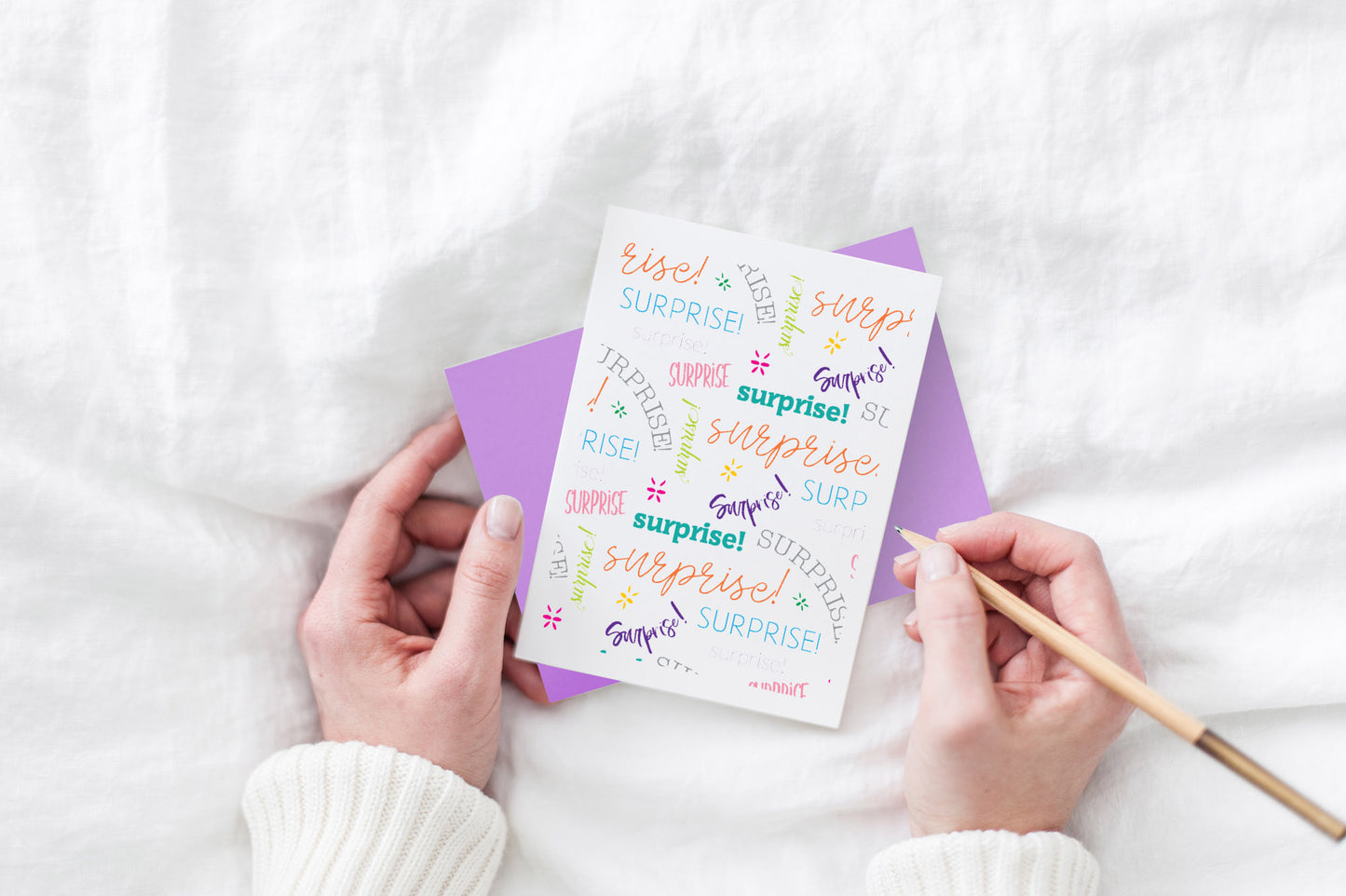 Greeting card and colorful envelope, seen from above, held by two hands along with a pencil, ready to write, against a white comforter.