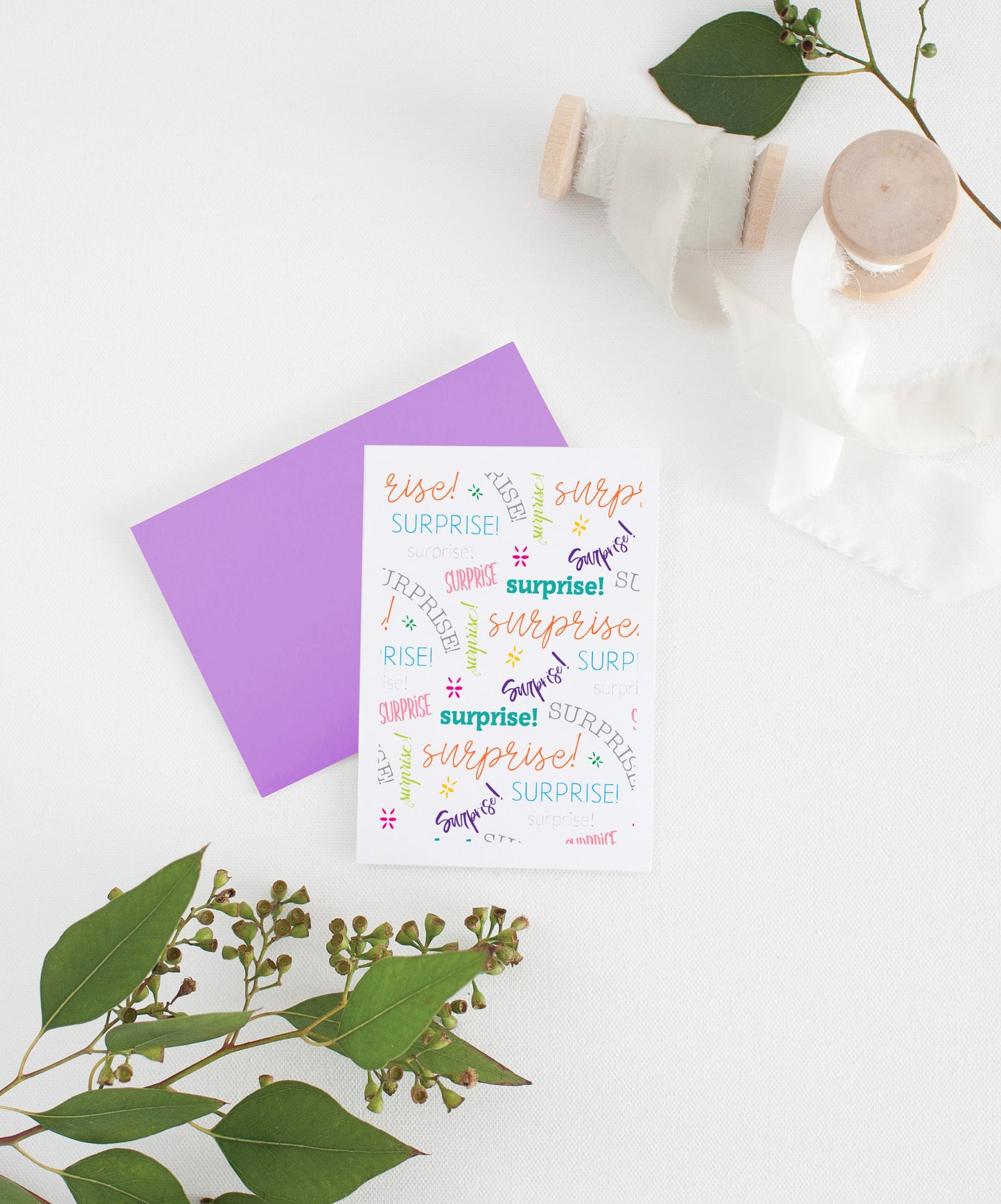 Greeting card with colorful envelope laying on a white surface with green leaves and cotton ribbon wound on wooden spools.