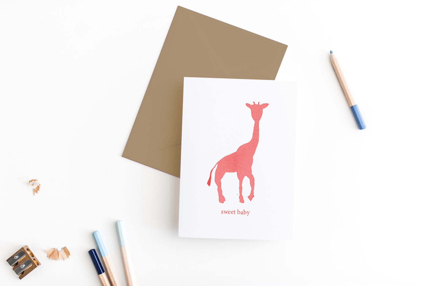 Greeting card, seen from above, with a kraft, square flap envelope, pencils, pencil sharpener and shavings on a white background.