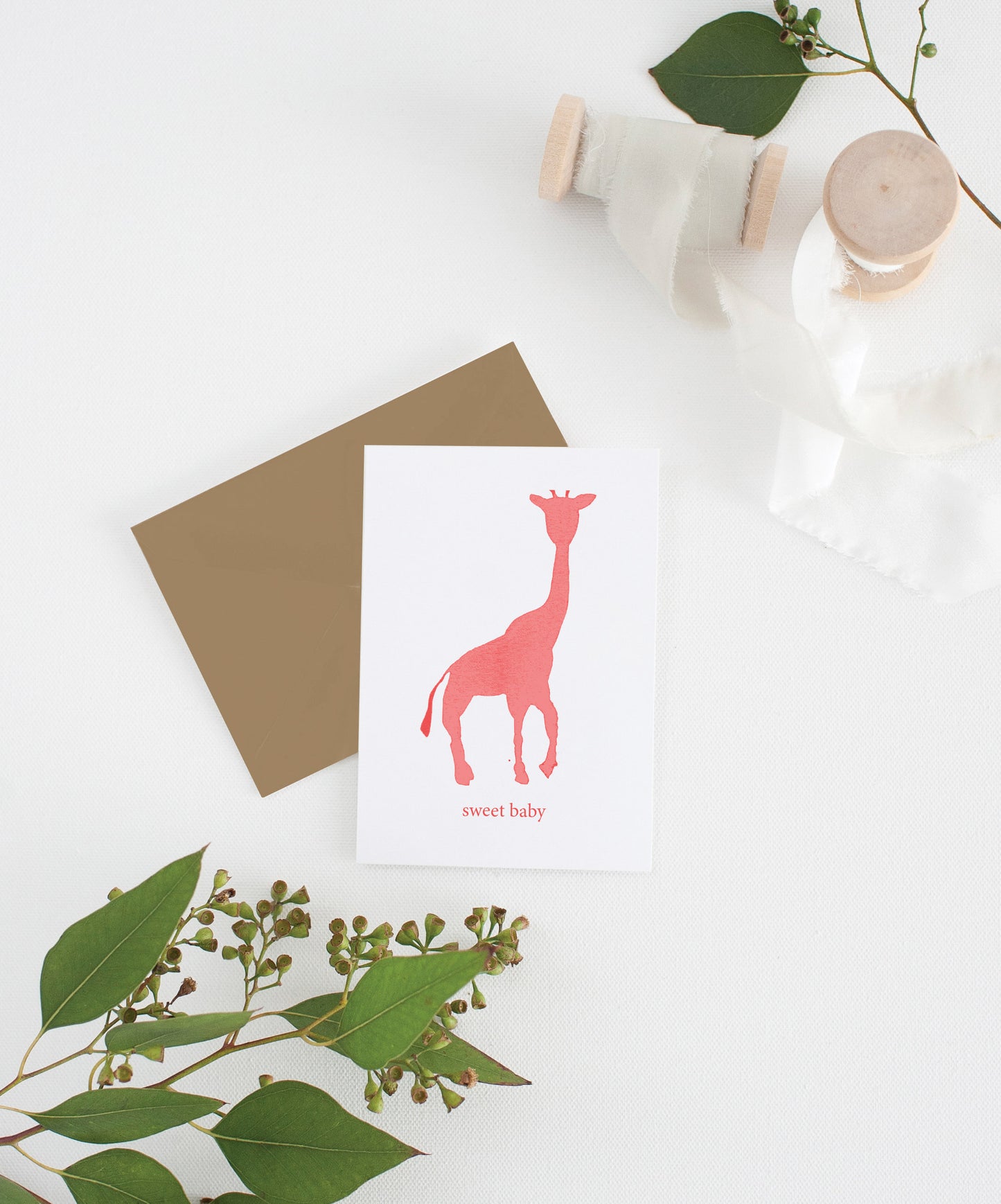 Greeting card with kraft envelope laying on a white surface with green leaves and cotton ribbon wound on wooden spools.
