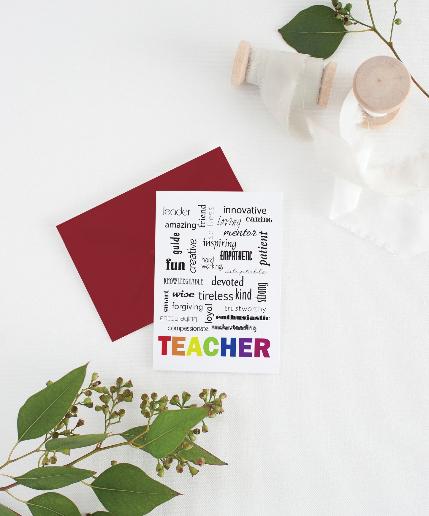 Greeting card with colorful envelope laying on a white surface with green leaves and cotton ribbon wound on wooden spools.