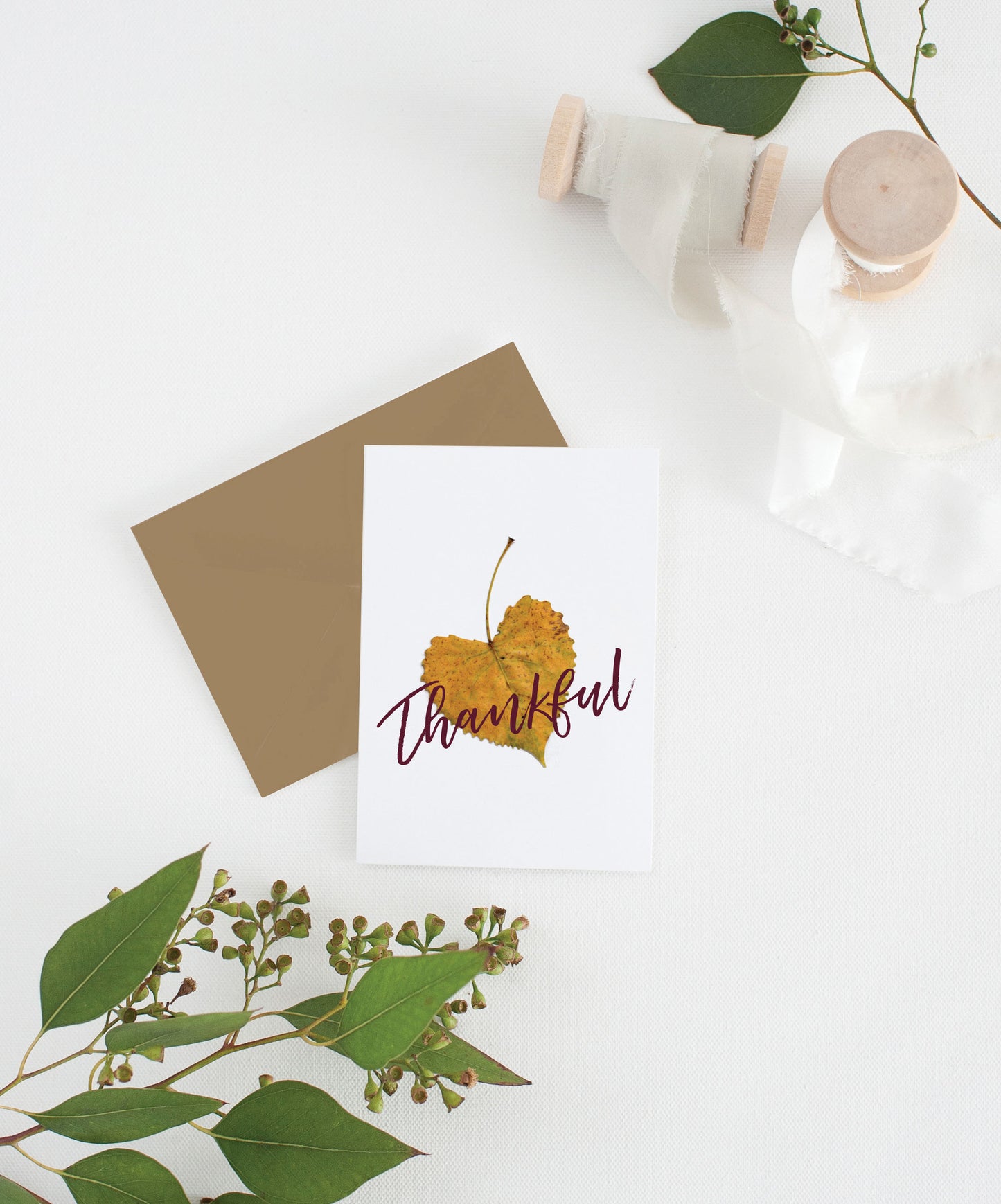 Greeting card with kraft envelope laying on a white surface with green leaves and cotton ribbon wound on wooden spools.