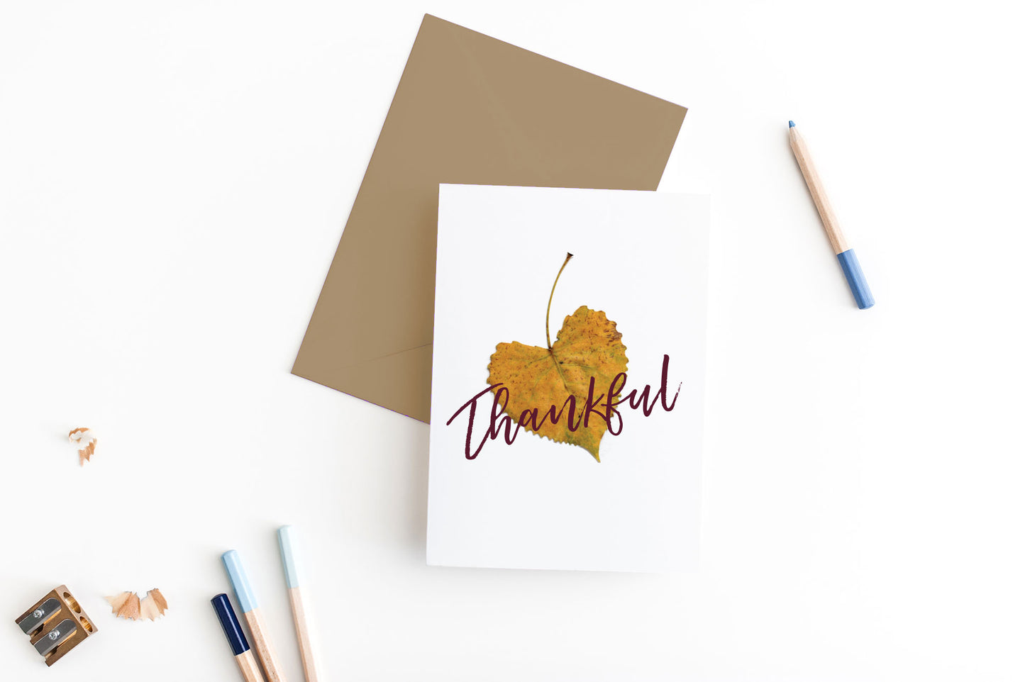 Greeting card, seen from above, with a kraft, square flap envelope, pencils, pencil sharpener and shavings on a white background.