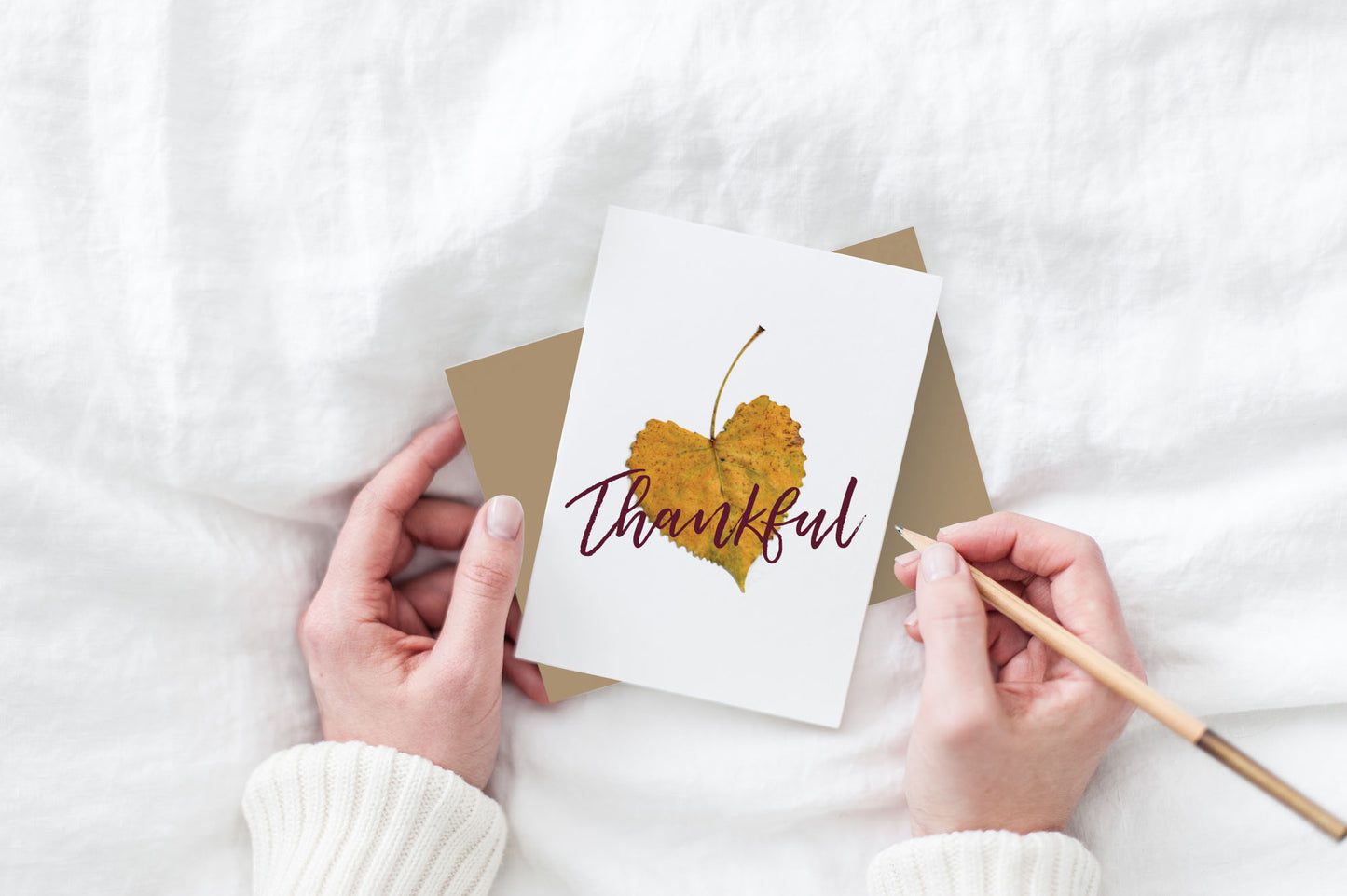 Greeting card and kraft envelope, seen from above, held by two hands along with a pencil, ready to write, against a white comforter.