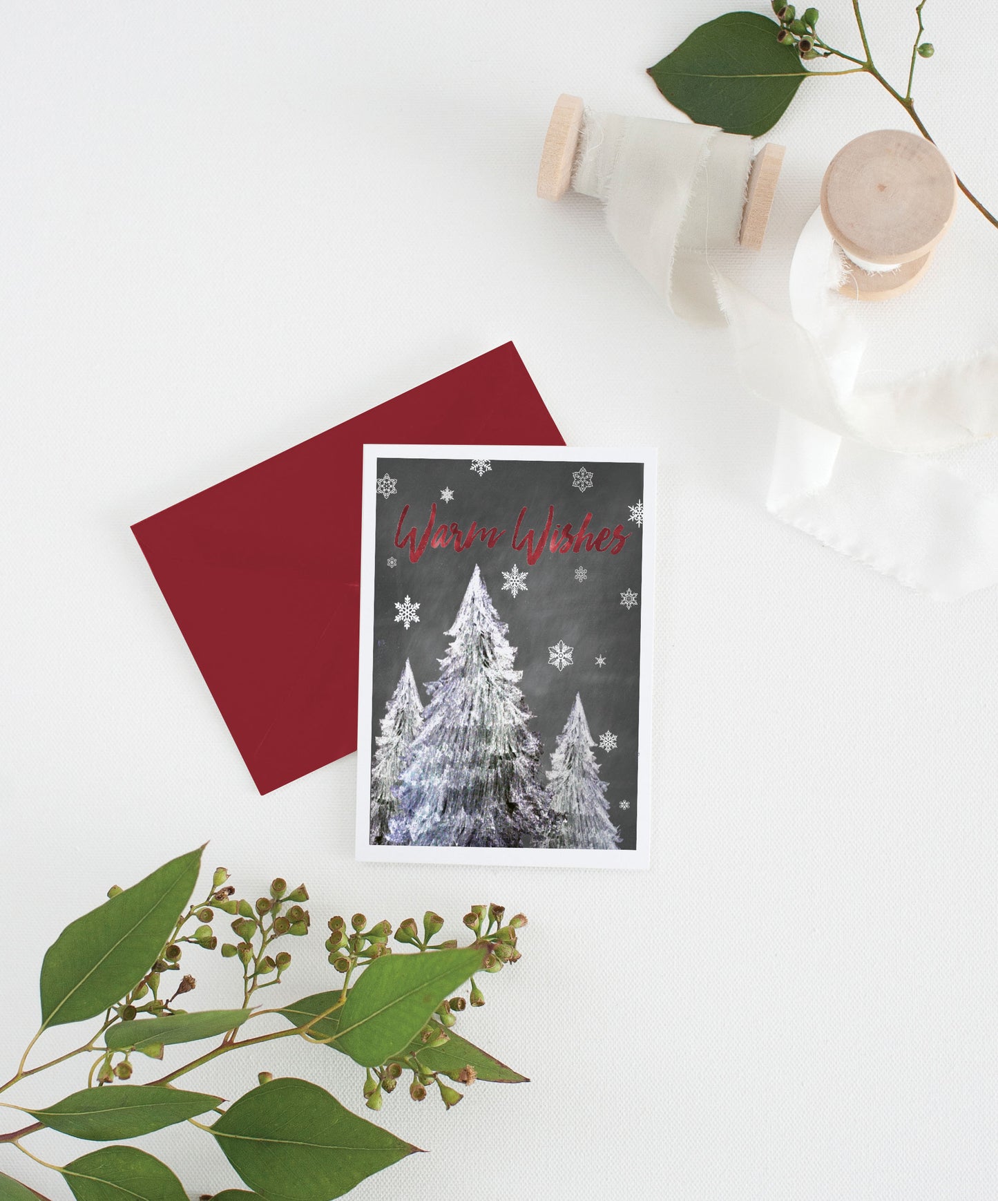 Greeting card with colorful envelope laying on a white surface with green leaves and cotton ribbon wound on wooden spools.