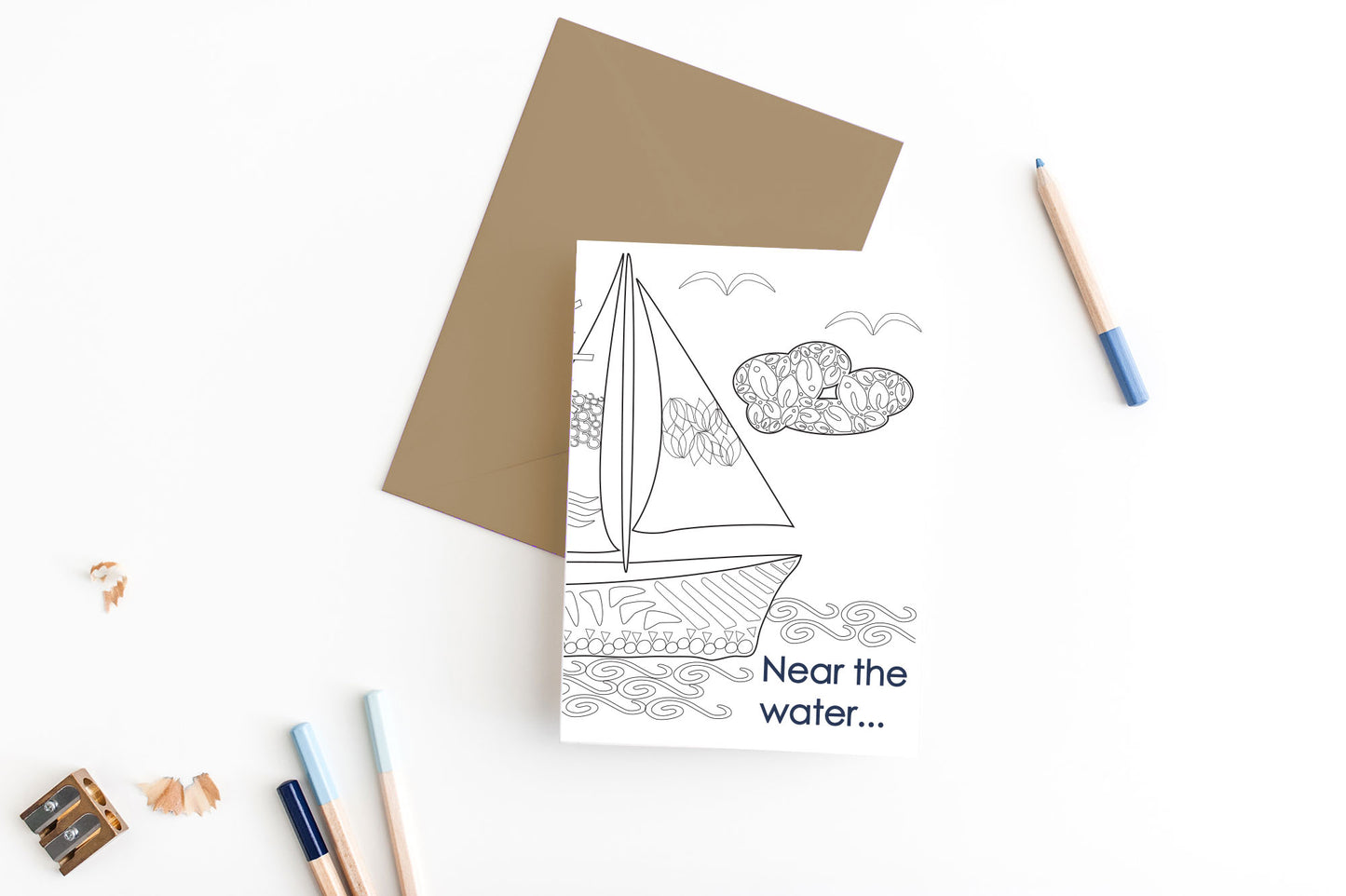 Greeting card, seen from above, with a kraft, square flap envelope, pencils, pencil sharpener and shavings on a white background.