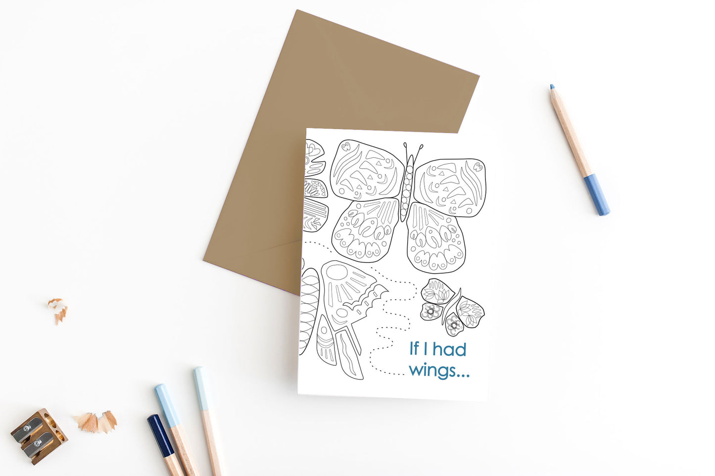 Greeting card, seen from above, with a kraft, square flap envelope, pencils, pencil sharpener and shavings on a white background.