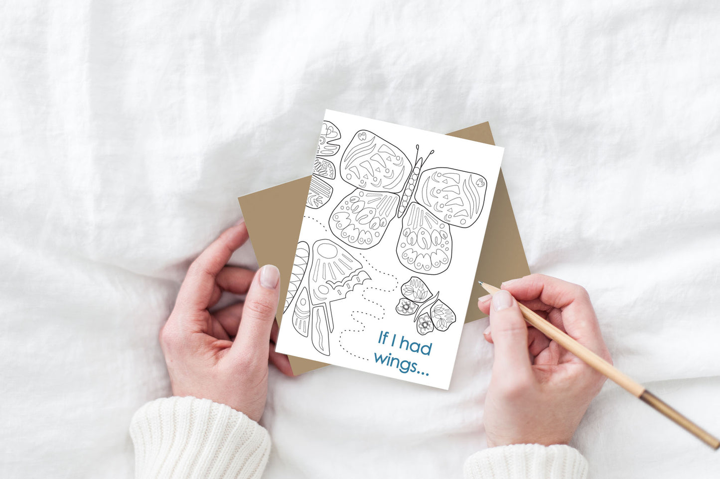 Greeting card and kraft envelope, seen from above, held by two hands along with a pencil, ready to write, against a white comforter.