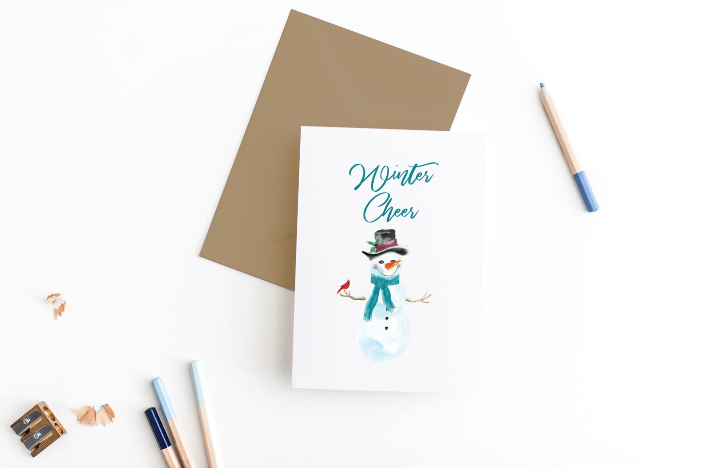 Greeting card, seen from above, with a kraft, square flap envelope, pencils, pencil sharpener and shavings on a white background.