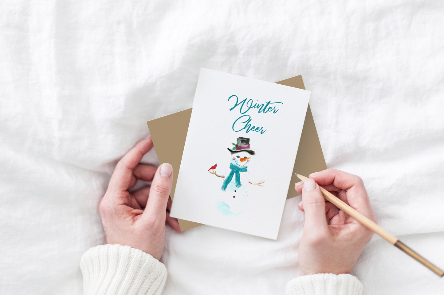Greeting card and kraft envelope, seen from above, held by two hands along with a pencil, ready to write, against a white comforter.