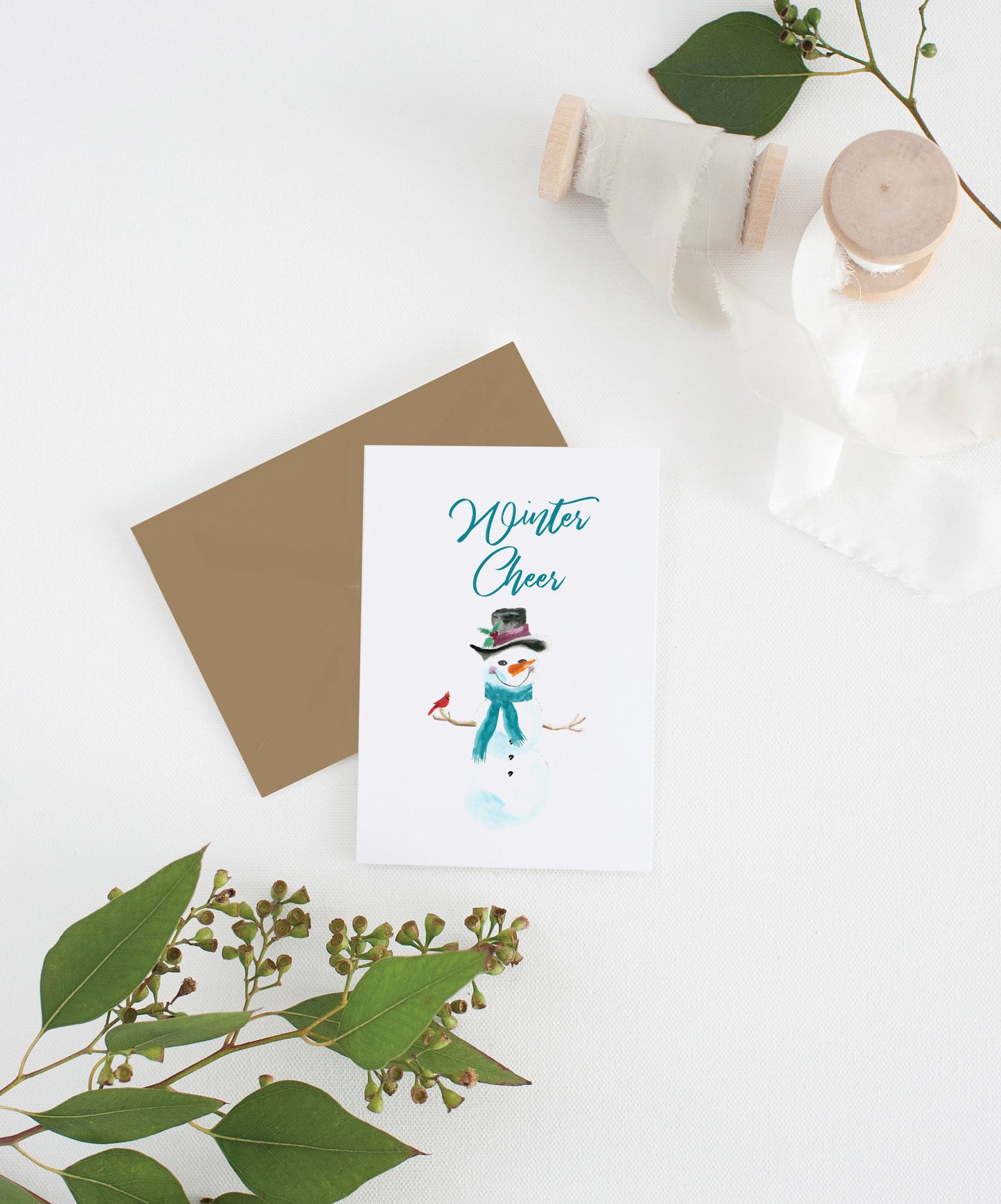 Greeting card with kraft envelope laying on a white surface with green leaves and cotton ribbon wound on wooden spools.