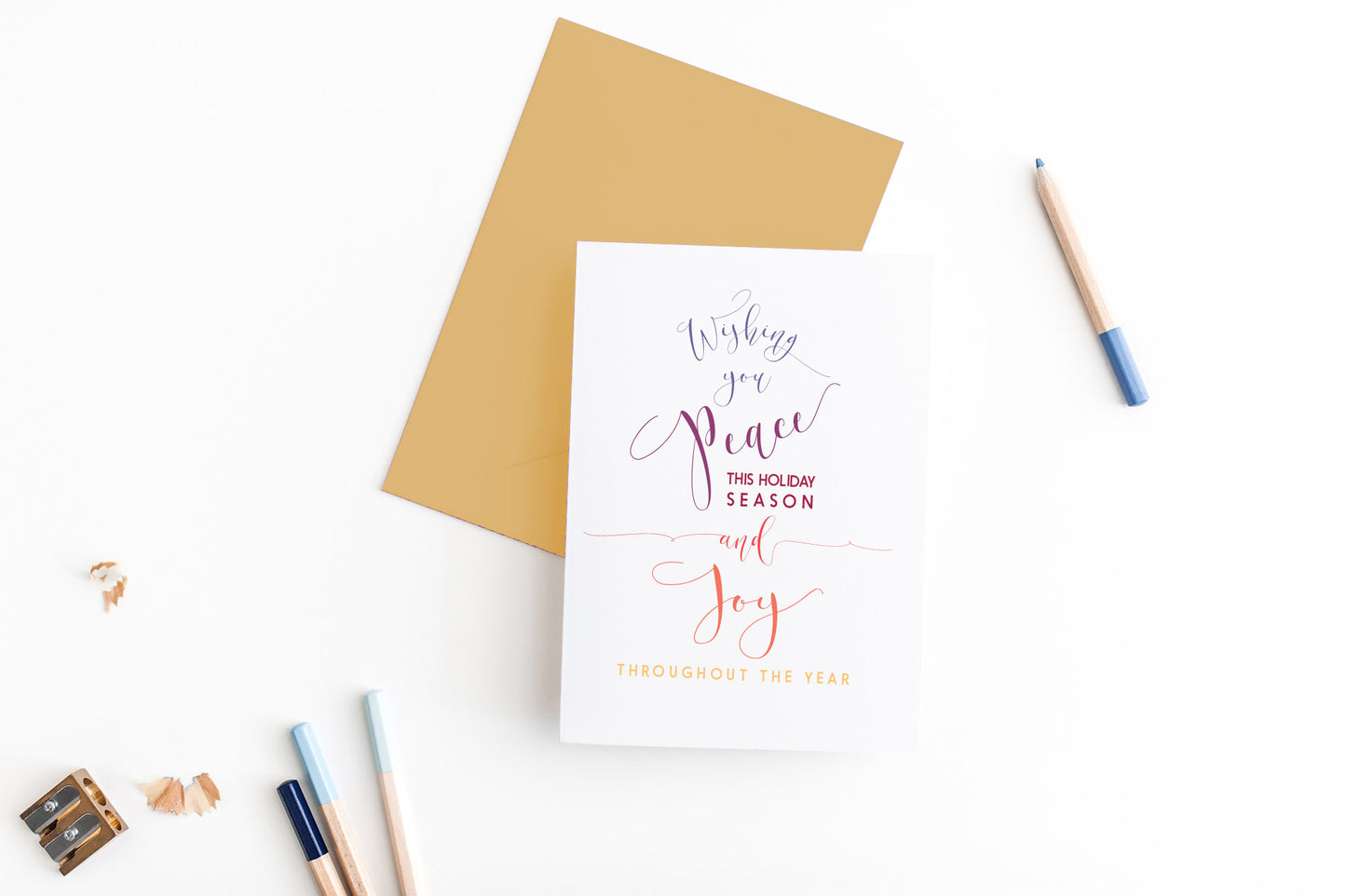 Greeting card, seen from above, with a colored, square flap envelope, pencils, pencil sharpener and shavings on a white background.