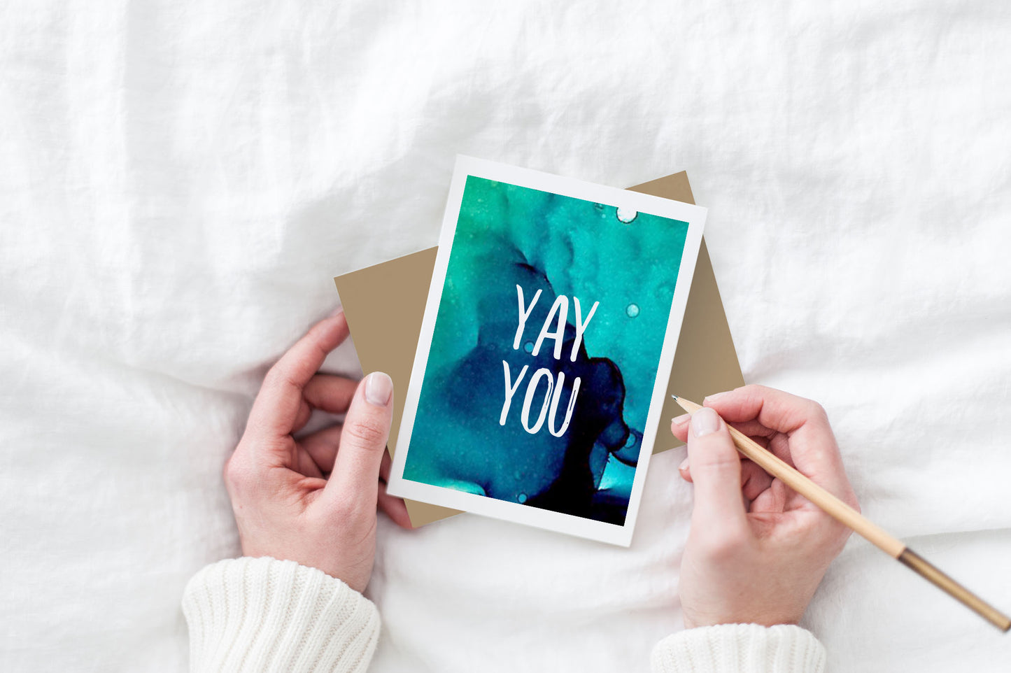 Greeting card and colorful envelope, seen from above, held by two hands along with a pencil, ready to write, against a white comforter.