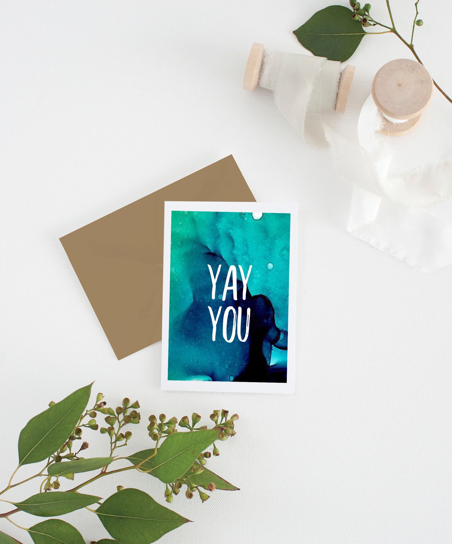 Greeting card with colorful envelope laying on a white surface with green leaves and cotton ribbon wound on wooden spools.