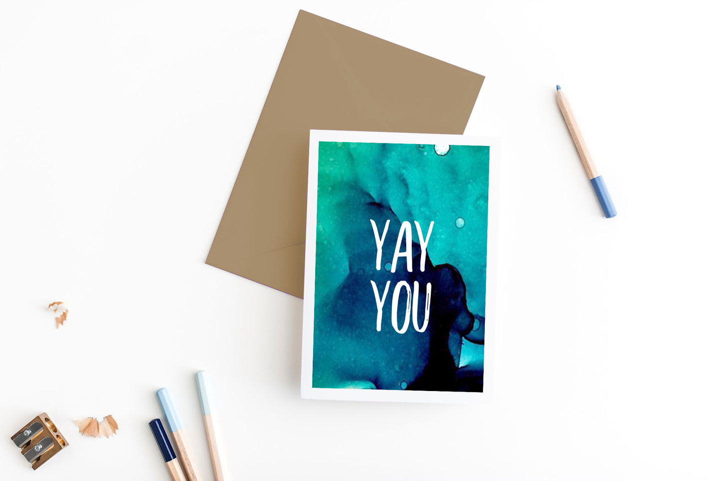Greeting card, seen from above, with a colored, square flap envelope, pencils, pencil sharpener and shavings on a white background.