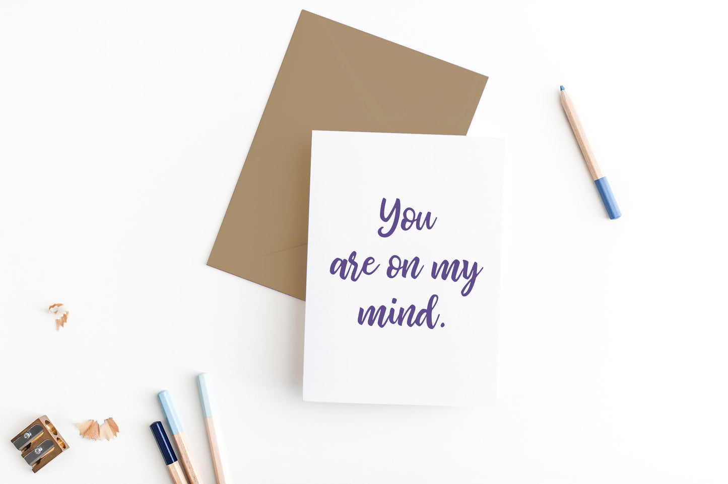 Greeting card, seen from above, with a colored, square flap envelope, pencils, pencil sharpener and shavings on a white background.