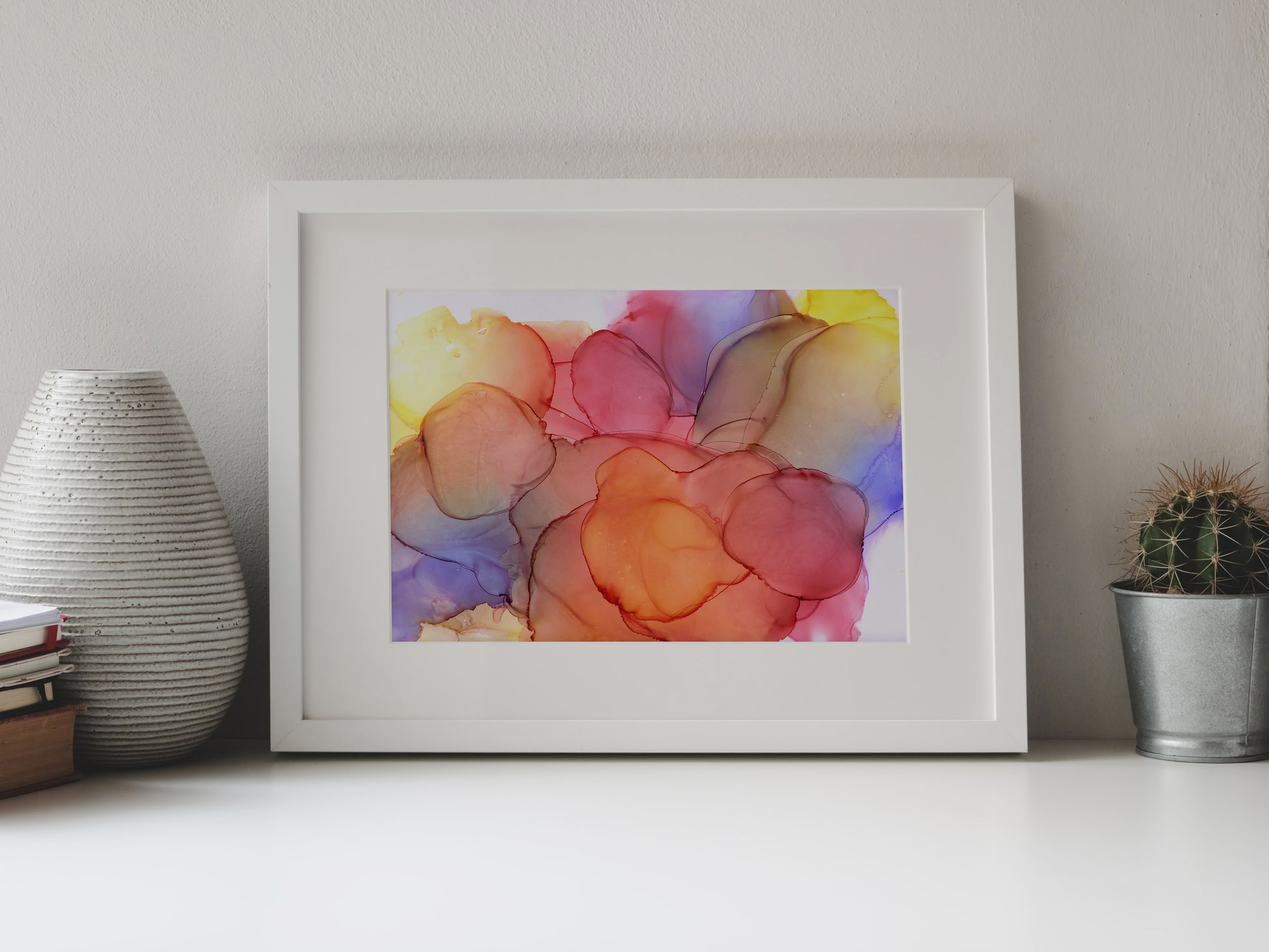 Digital mockup of Sundrenched Glow framed in white on a white table top, leaning up against a white background, surrounded by a vase, stack of books, and tiny cactus in a pail.