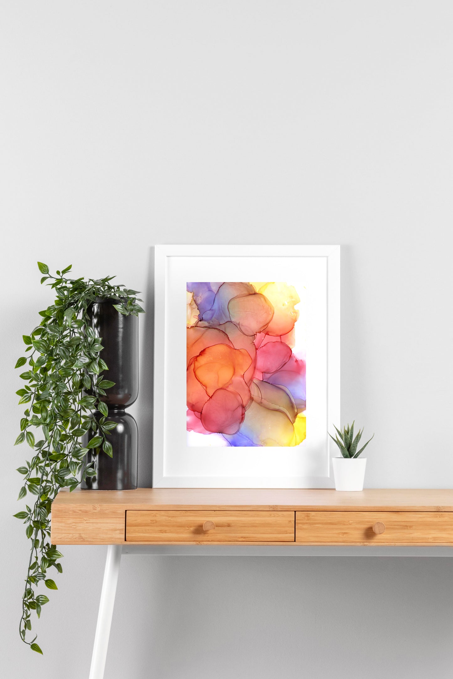 Digital mockup of Sundrenched Glow framed in white on a wooden desk, leaning up against a neutral wall with a small succulent in a white planter on one side and greenery in a taller black planter on the other.