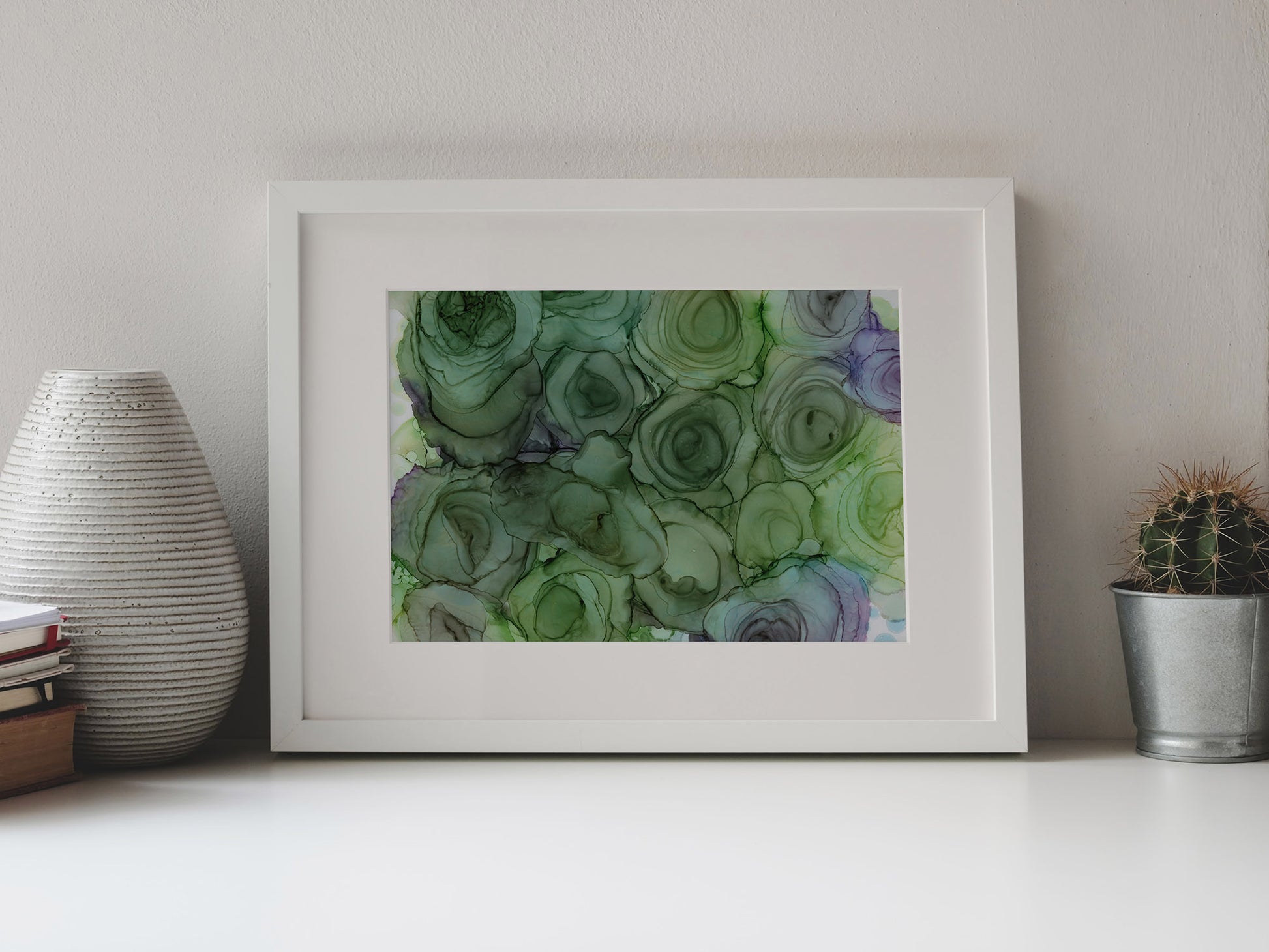 Digital mockup of Verdant Blooms framed in white on a white table top, leaning up against a white background, surrounded by a vase, stack of books, and tiny cactus in a pail.