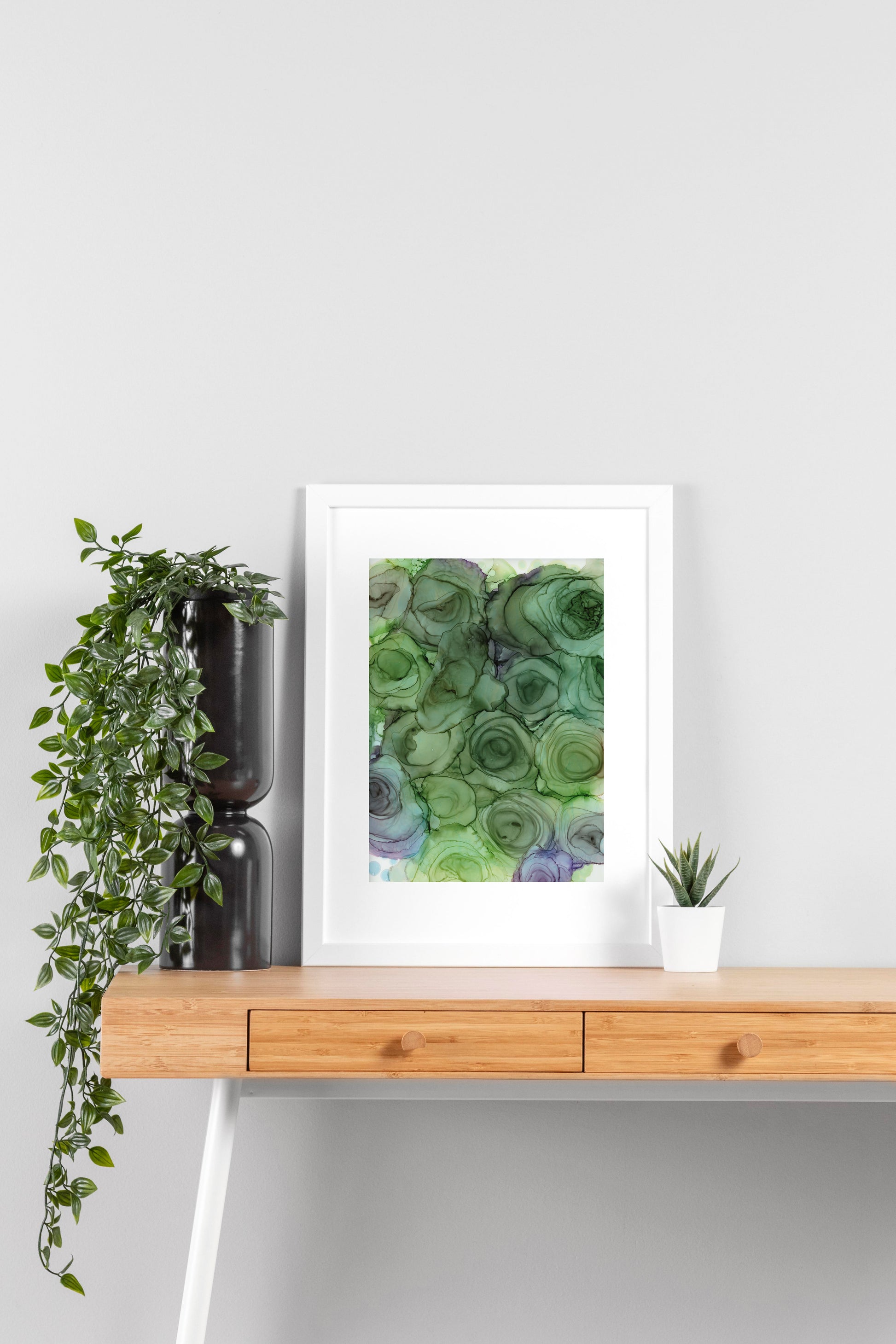 Digital mockup of Verdant Blooms framed in white on a wooden desk, leaning up against a neutral wall with a small succulent in a white planter on one side and greenery in a taller black planter on the other.