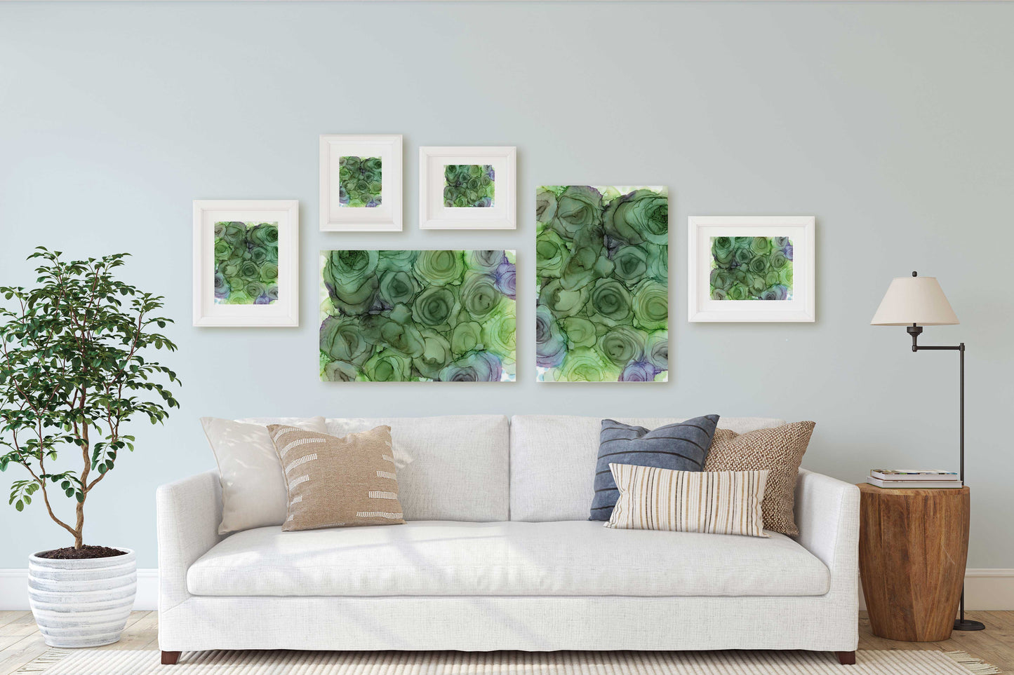 A styled living room scene showing a white sofa with neutral and striped pillows, a potted plant, a floor lamp, and a set of six framed green abstract art prints displayed on a light gray wall.