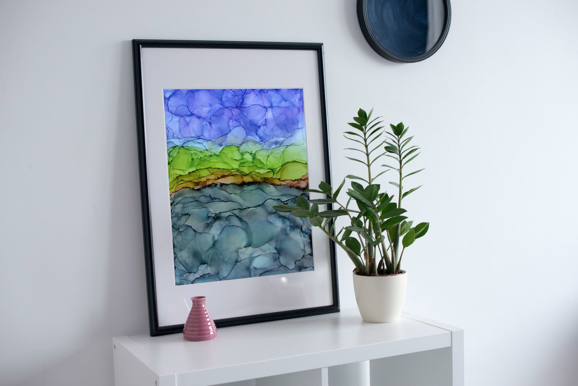 Zoomed out digital mockup of Break in the Clouds framed in black leaning on a white shelf against a white wall next to a small white planter with a green multi stem plant.