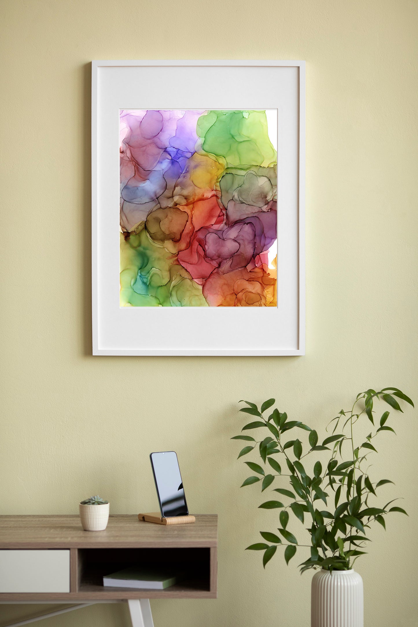 Digital mockup of a living room with Surrounded By Abundance framed in white hung on a pale yellow wall with a short wooden table and greenery in a cream vase below. 