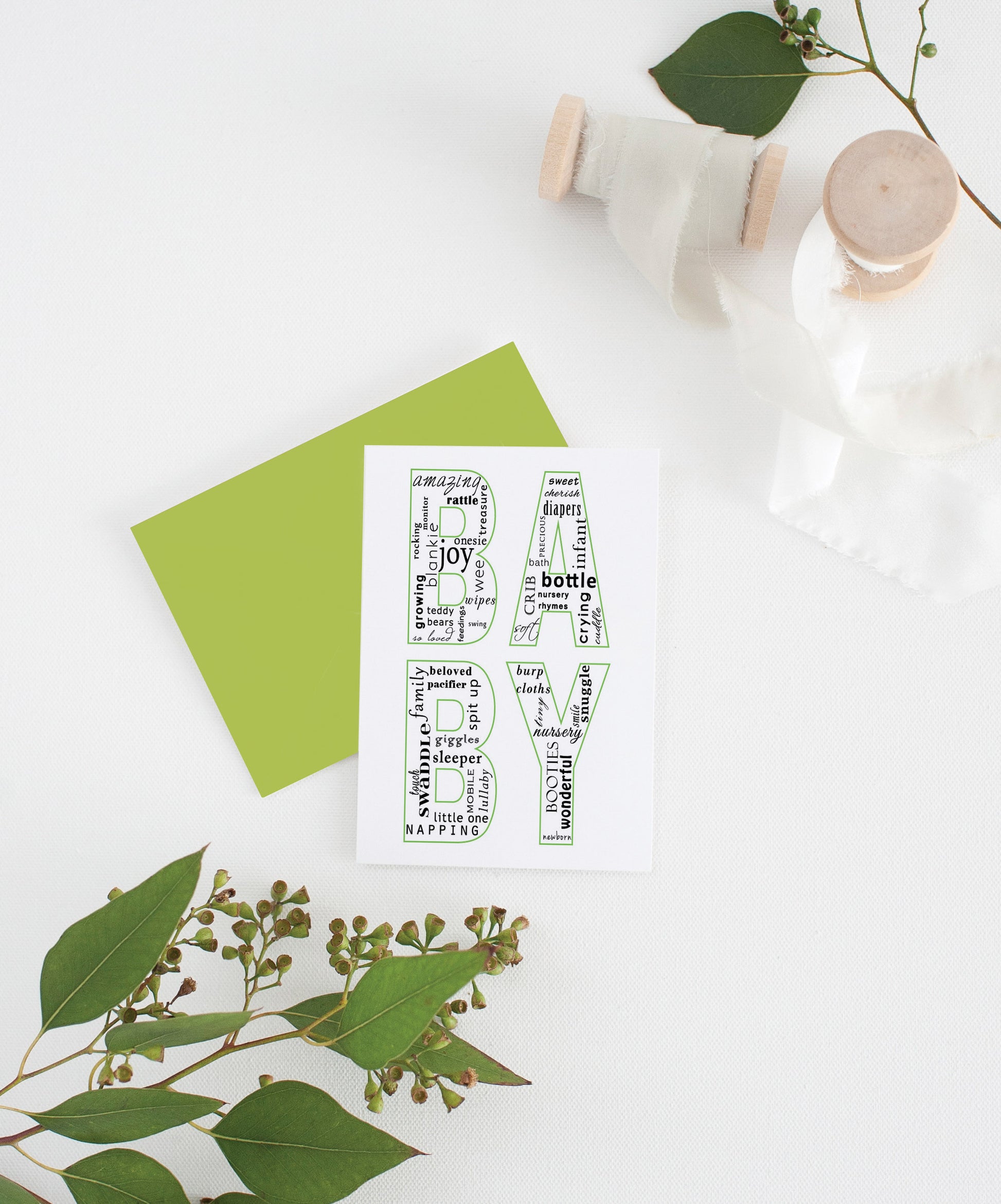 Greeting card with colorful envelope laying on a white surface with green leaves and cotton ribbon wound on wooden spools.