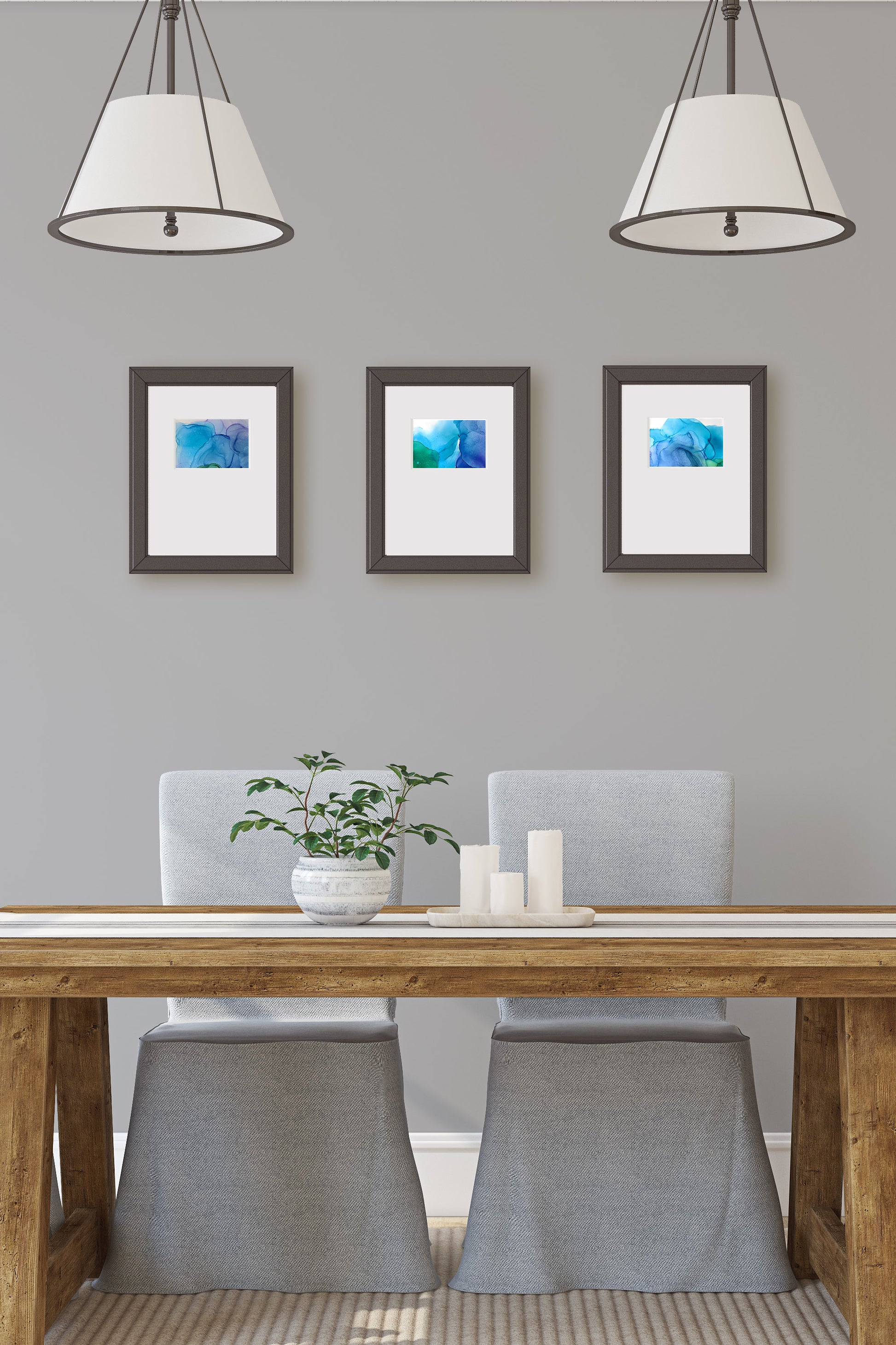 Digital mockup of the three paintings framed in black hung on a grey wall above a farmhouse style wooden dining table.  Two light fixtures hang from above and a ceramic planter and pillar candles sit on a fabric table runner in front of fabric covered chairs.