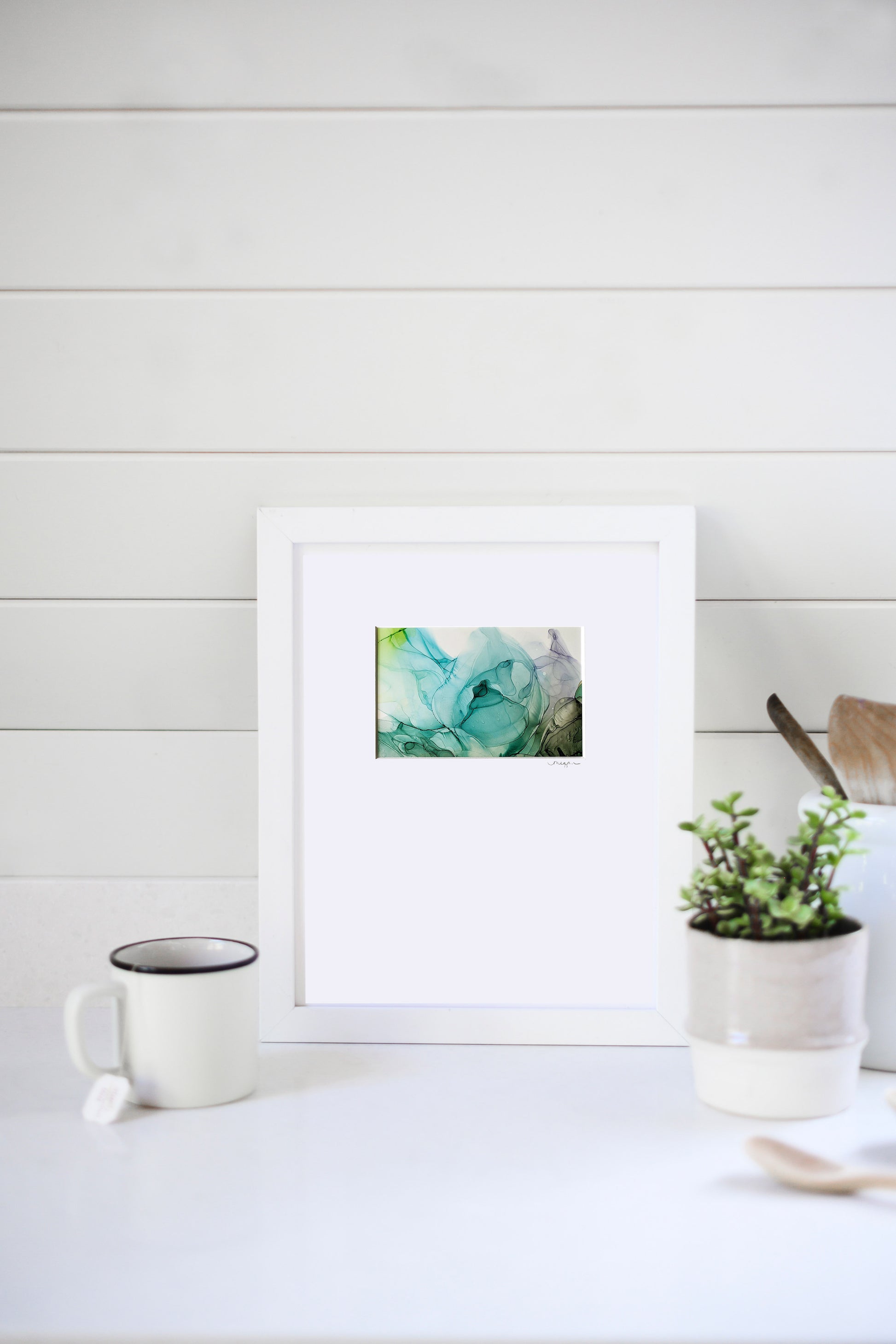 Digital mockup of the painting framed in white leaning against a horizontal white beadboard wall.  A ceramic mug, ceramic planter with greenery, and wooden utensils sit to the sides.