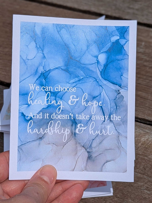 Card with blue abstract ink painting design and motivational wording held by a hand on a wooden surface.