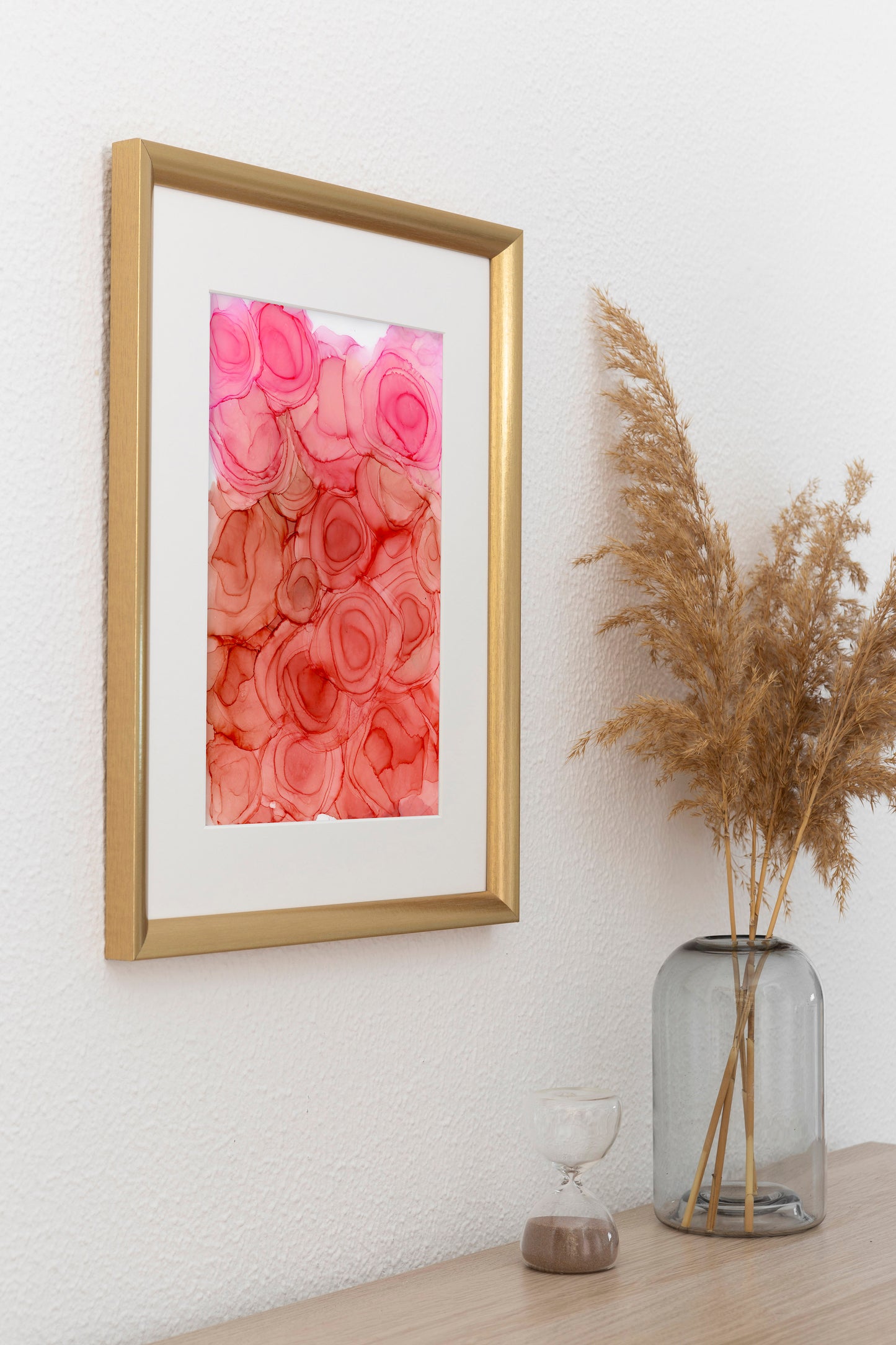 Digital mockup of I am Blooming framed in gold hung on a white wall.  A glass vase with dried grasse and a glass sand timer sit on the table below.