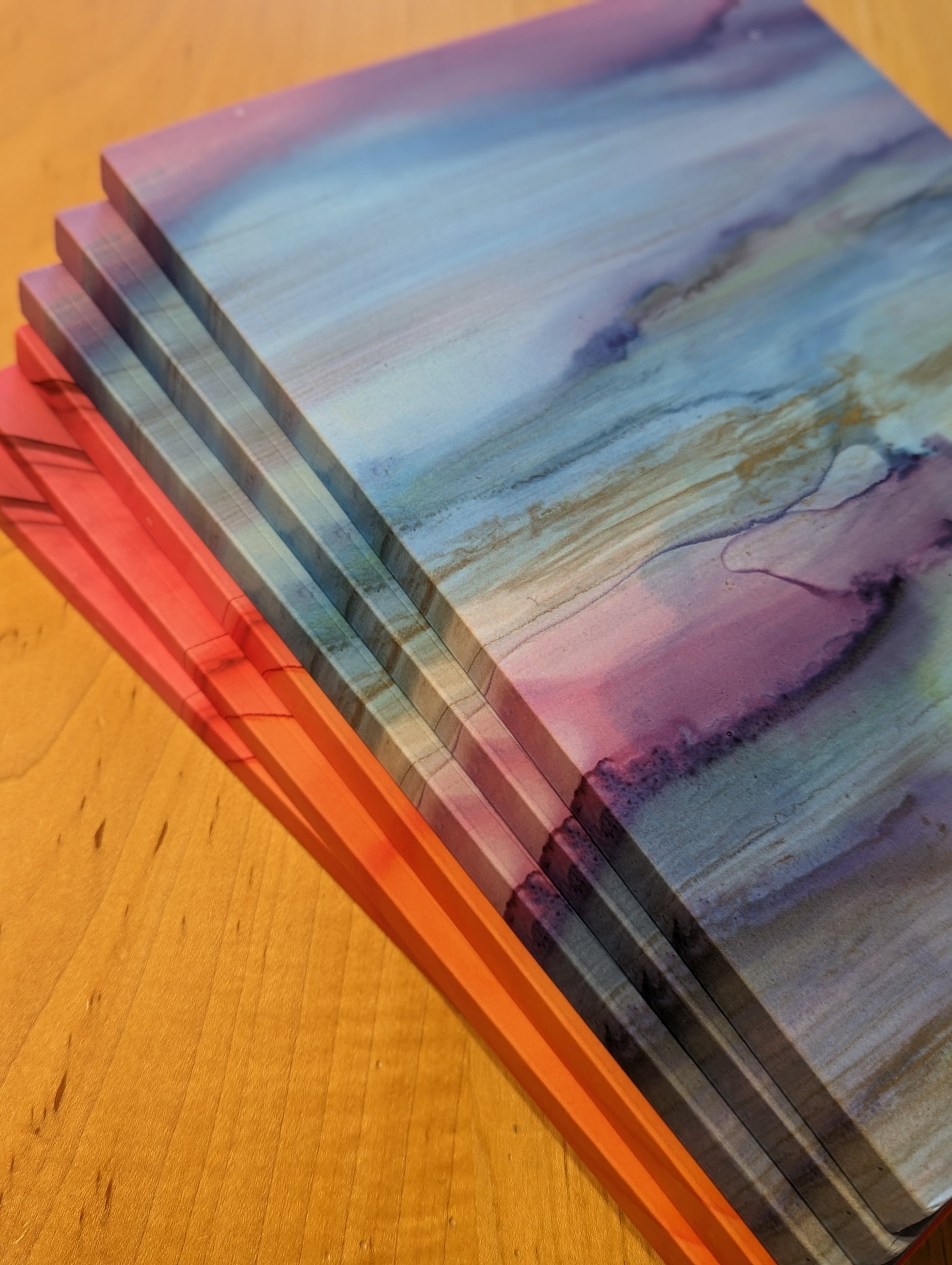 Stack of three notebooks showing the cover style Sundrenched Glow and three notebooks showing the cover style Pirr sitting on a wood table.