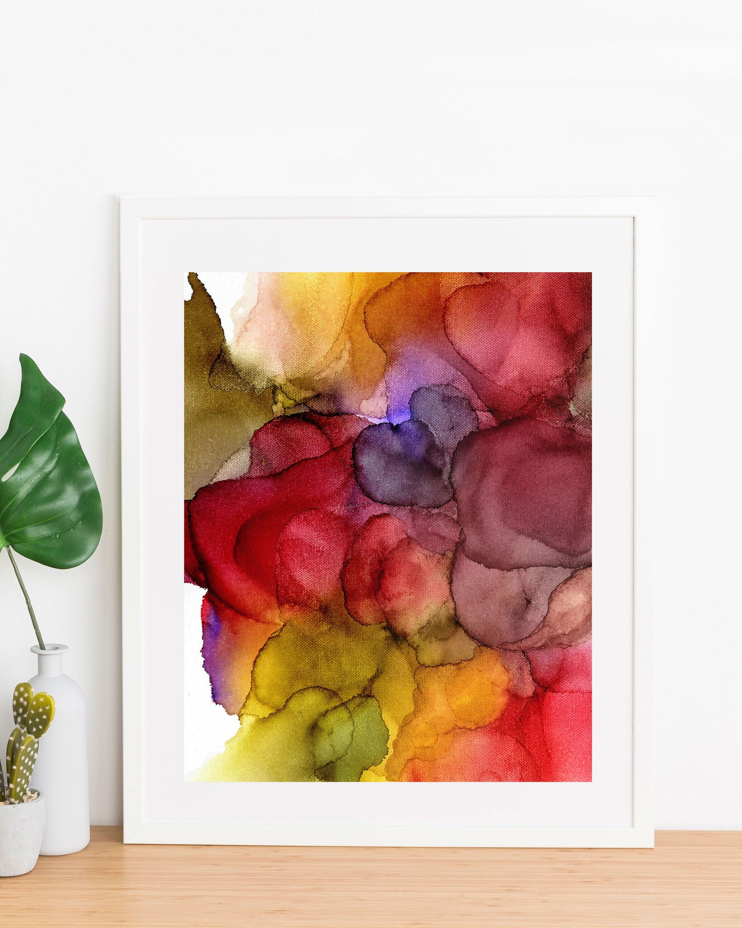 Abundance painting displayed in a white frame propped up against a white wall, on a wood table with a small vase and planter on the left.