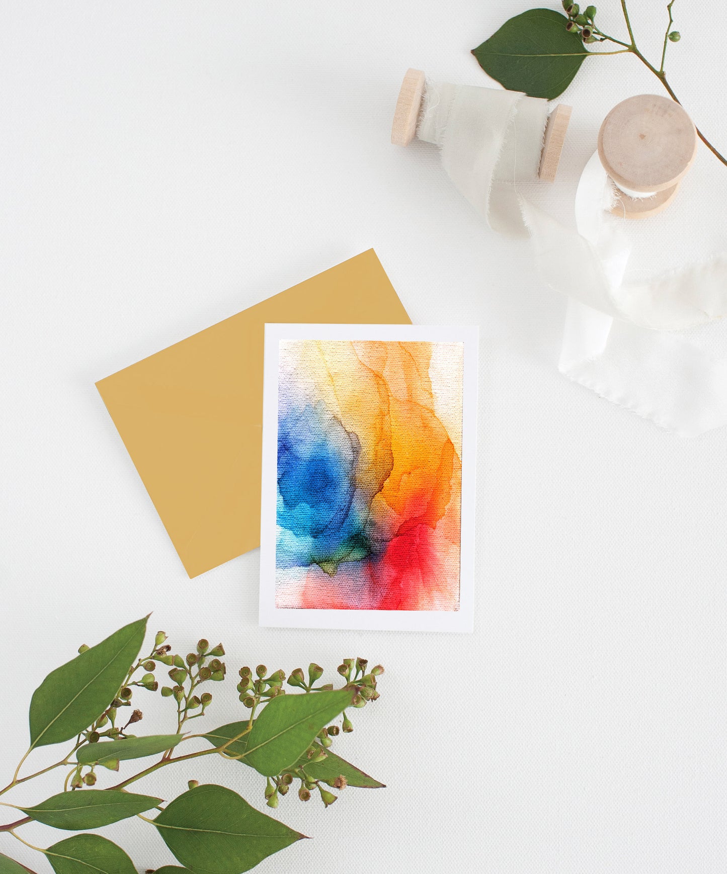 Greeting card with colorful envelope laying on a white surface with green leaves and cotton ribbon wound on wooden spools.