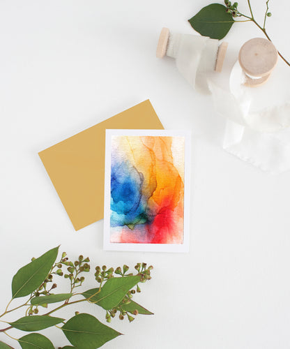 Greeting card with colorful envelope laying on a white surface with green leaves and cotton ribbon wound on wooden spools.