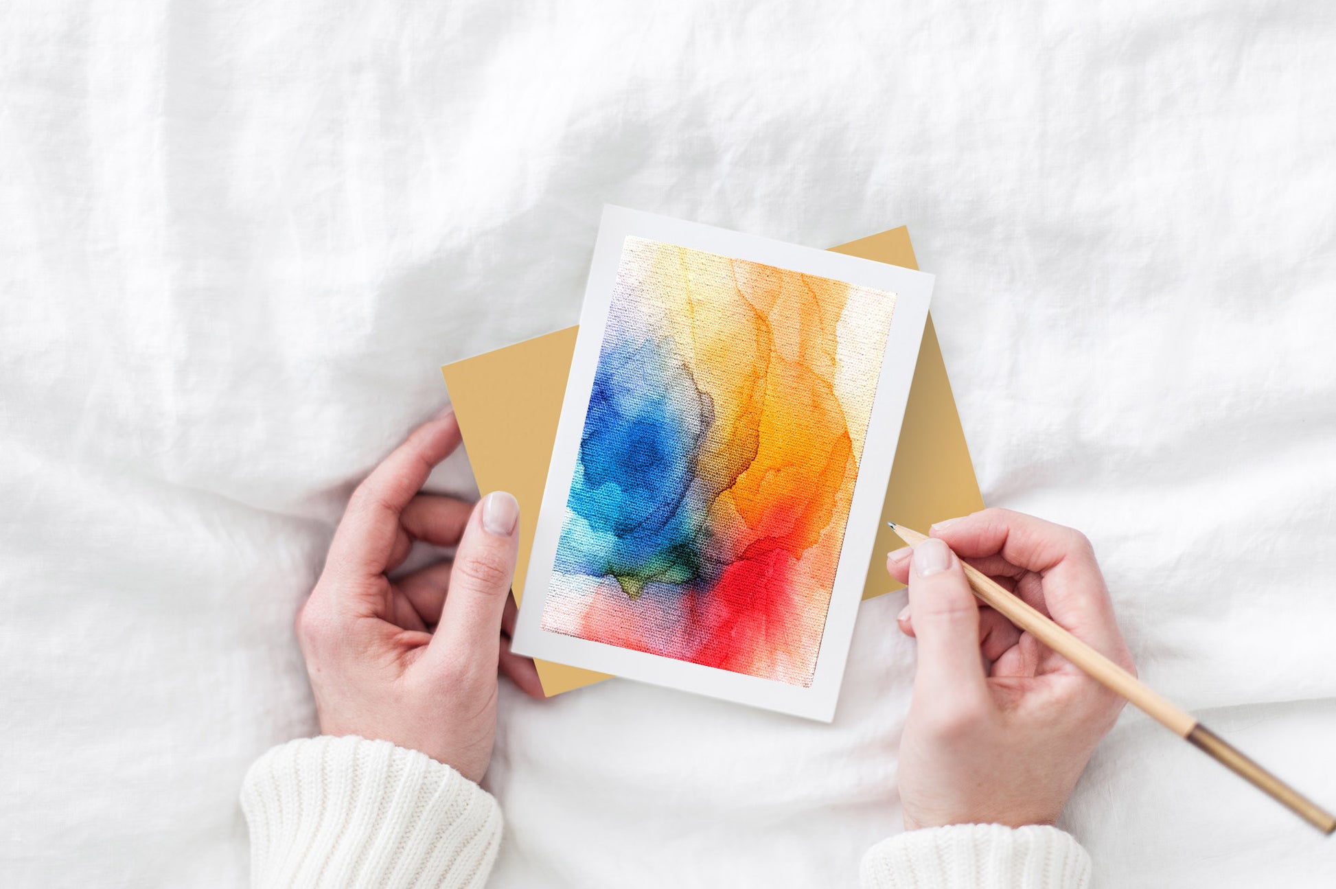 Greeting card and colorful envelope, seen from above, held by two hands along with a pencil, ready to write, against a white comforter.