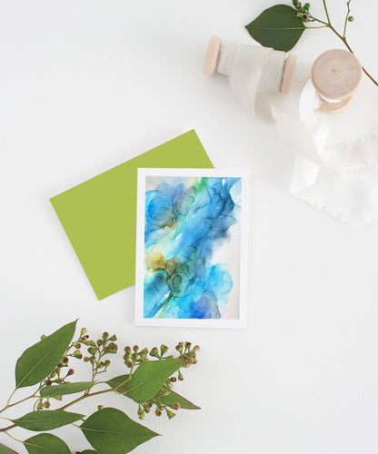 Greeting card with colorful envelope laying on a white surface with green leaves and cotton ribbon wound on wooden spools.