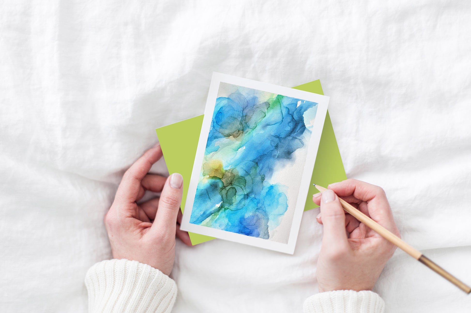 Greeting card and colorful envelope, seen from above, held by two hands along with a pencil, ready to write, against a white comforter.