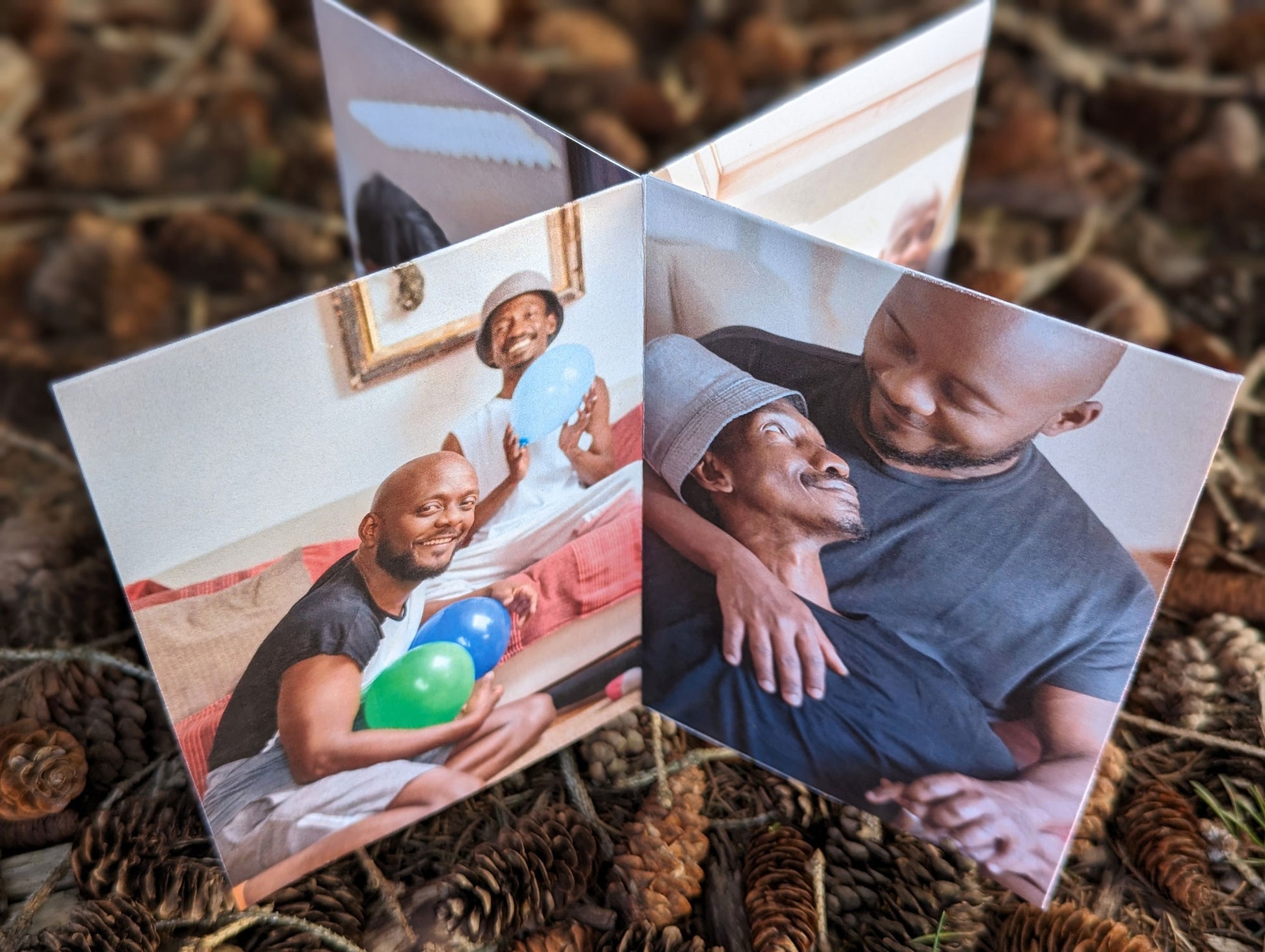 Centerpiece sitting on a bed of small pinecones seen from overhead with photos of an LGBTQ+ couple smiling at each other and one where they are holding colorful balloons and smiling at the camera.