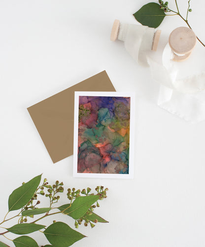 Greeting card with colorful envelope laying on a white surface with green leaves and cotton ribbon wound on wooden spools.