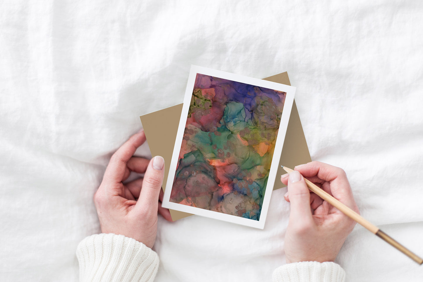 Greeting card and colorful envelope, seen from above, held by two hands along with a pencil, ready to write, against a white comforter.