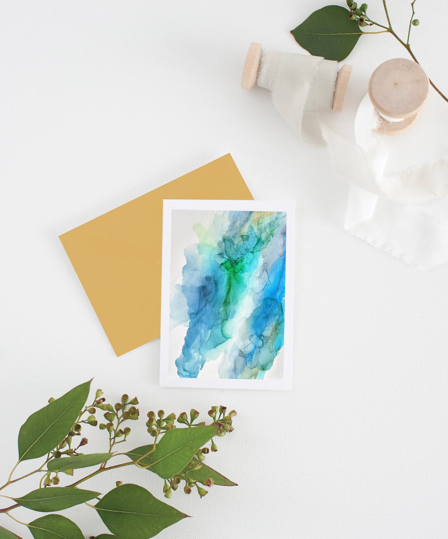 Greeting card with colorful envelope laying on a white surface with green leaves and cotton ribbon wound on wooden spools.