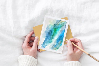 Greeting card and colorful envelope, seen from above, held by two hands along with a pencil, ready to write, against a white comforter.