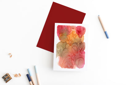 Greeting card and colorful envelope, seen from above, held by two hands along with a pencil, ready to write, against a white comforter.