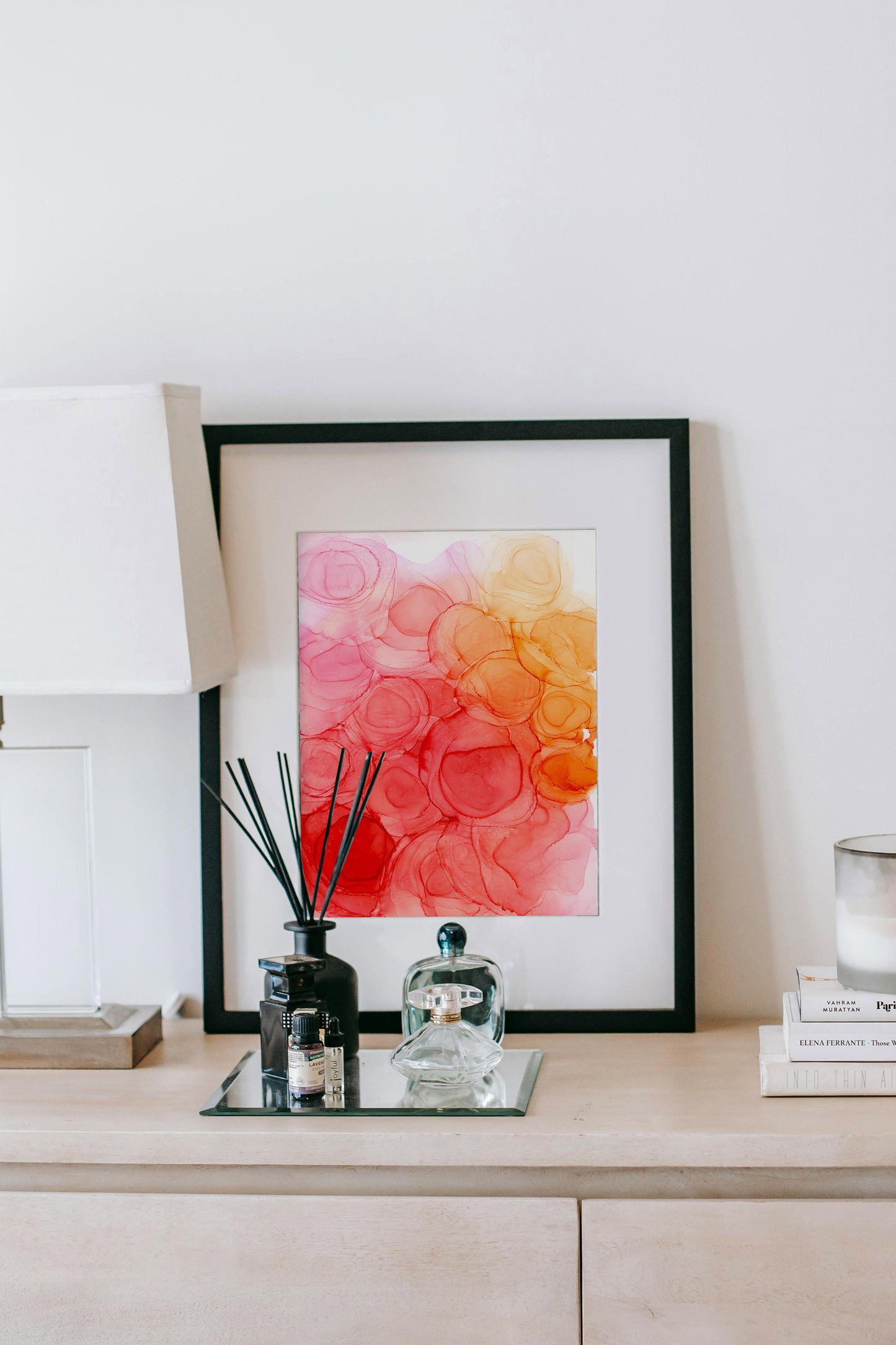 Digital mockup of a living room with Energizing Spring framed in black, propped up on a sideboard, against a white wall along with a lamp, incense, perfume bottles, a stack of books and a candle.