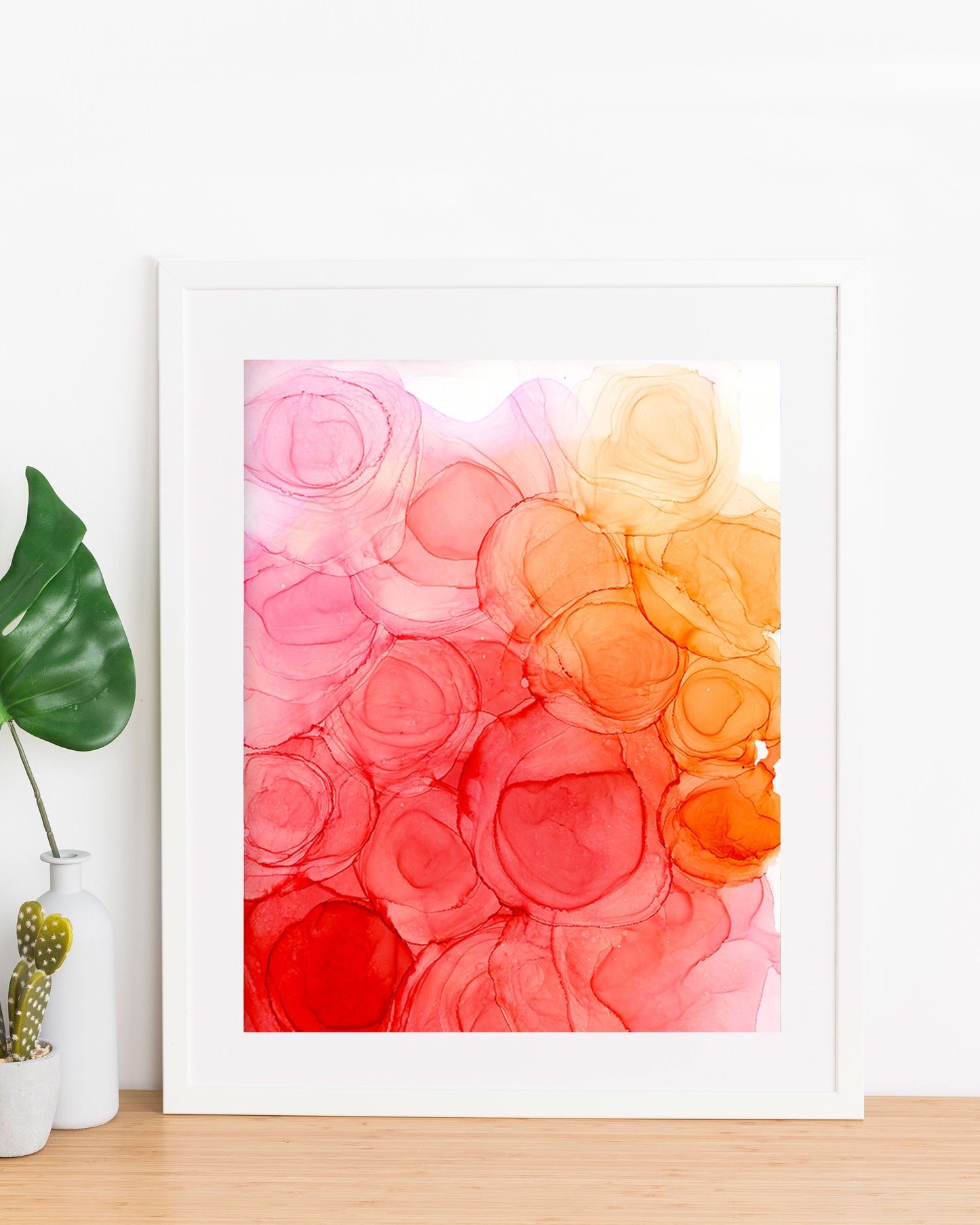 Energizing Spring painting displayed in a white frame propped up against a white wall, on a wood table with a small vase and planter on the left.