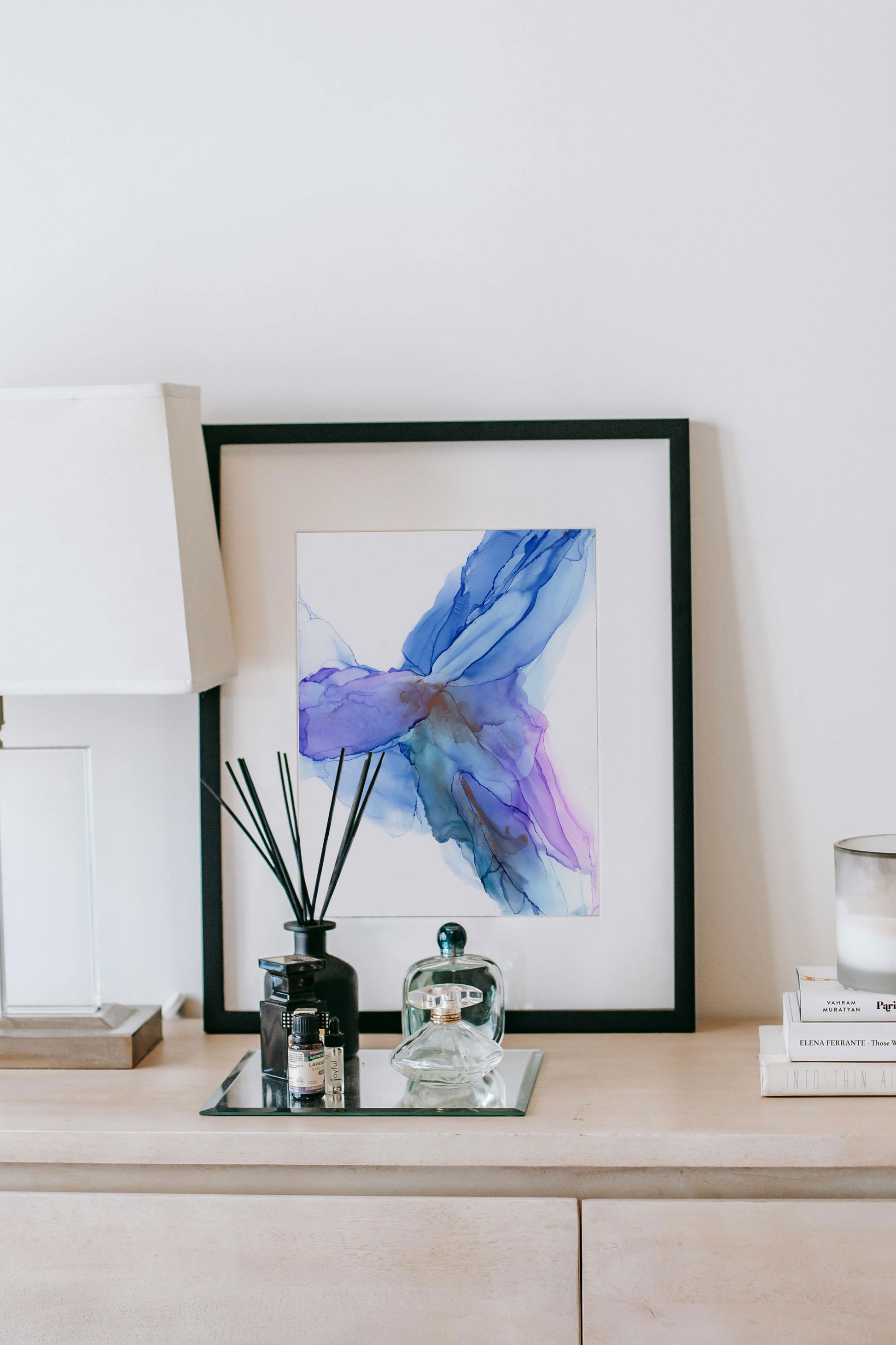 Digital mockup of a living room with Graceful Movement framed in black, propped up on a sideboard, against a white wall along with a lamp, incense, perfume bottles, a stack of books and a candle.